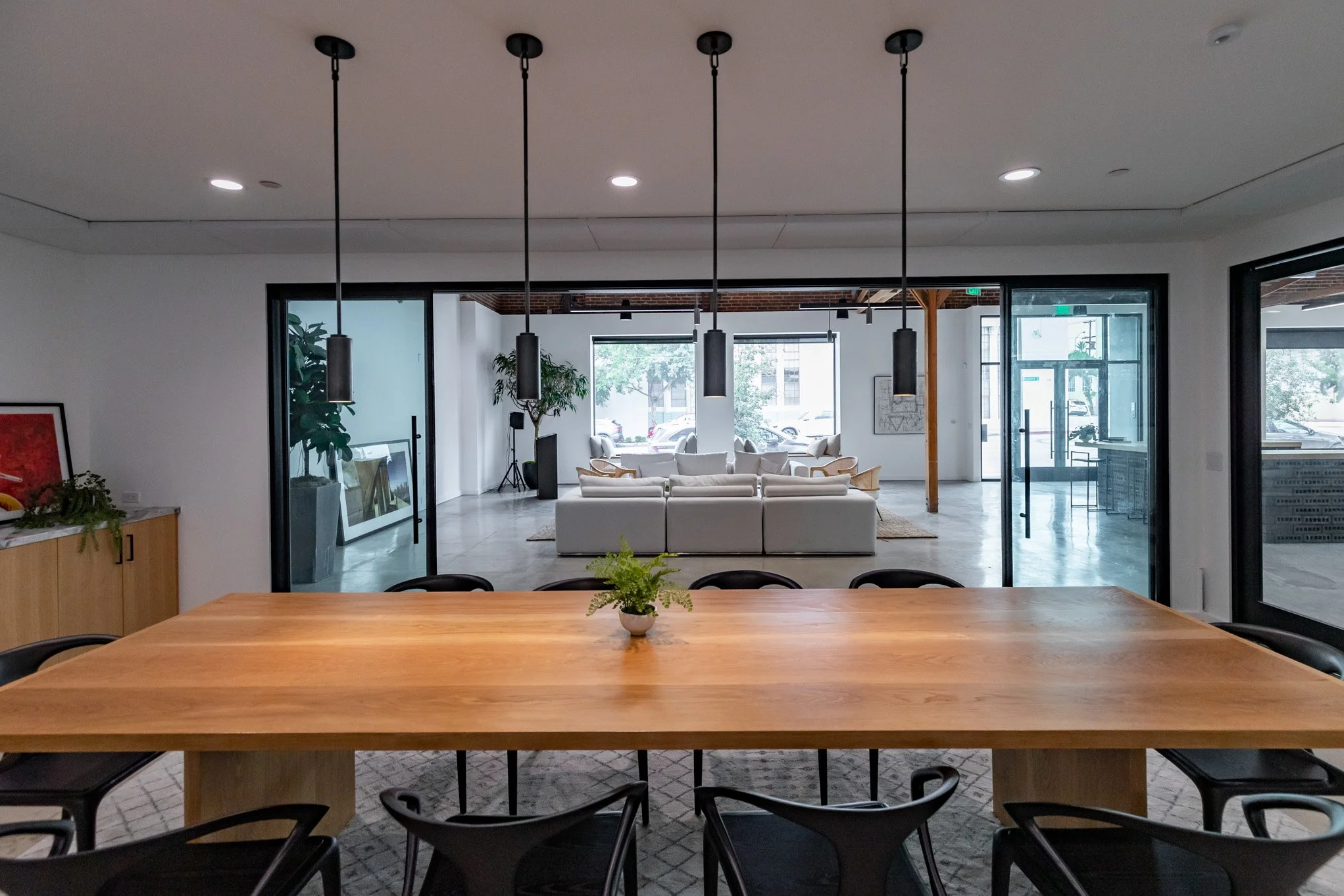 Modern conference room with a large wooden table, black chairs, and a living room visible in the background with white sofas, plants, and large windows.