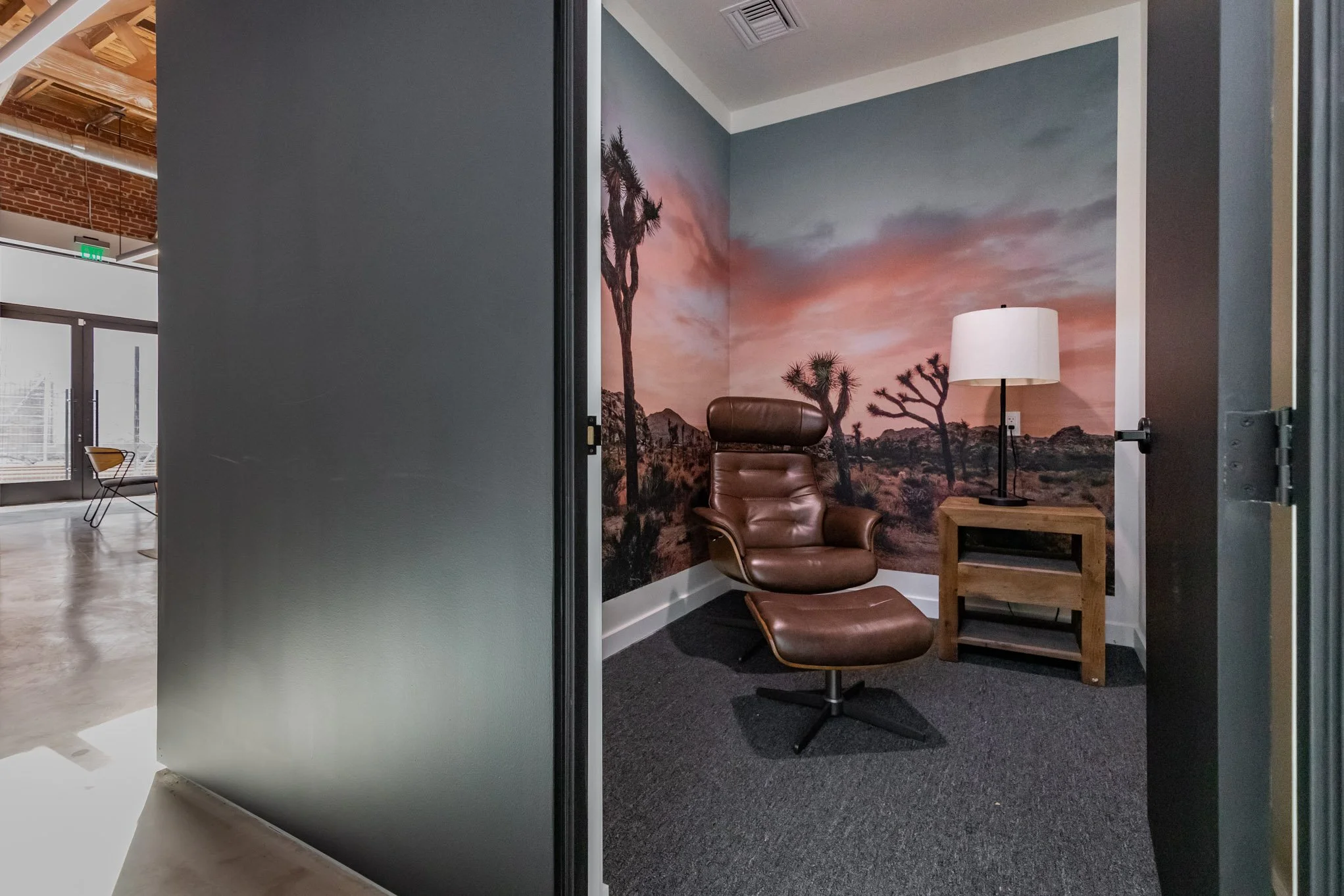 A small office nook with a leather chair, a wooden side table with a lamp, and a desert landscape mural on the wall.