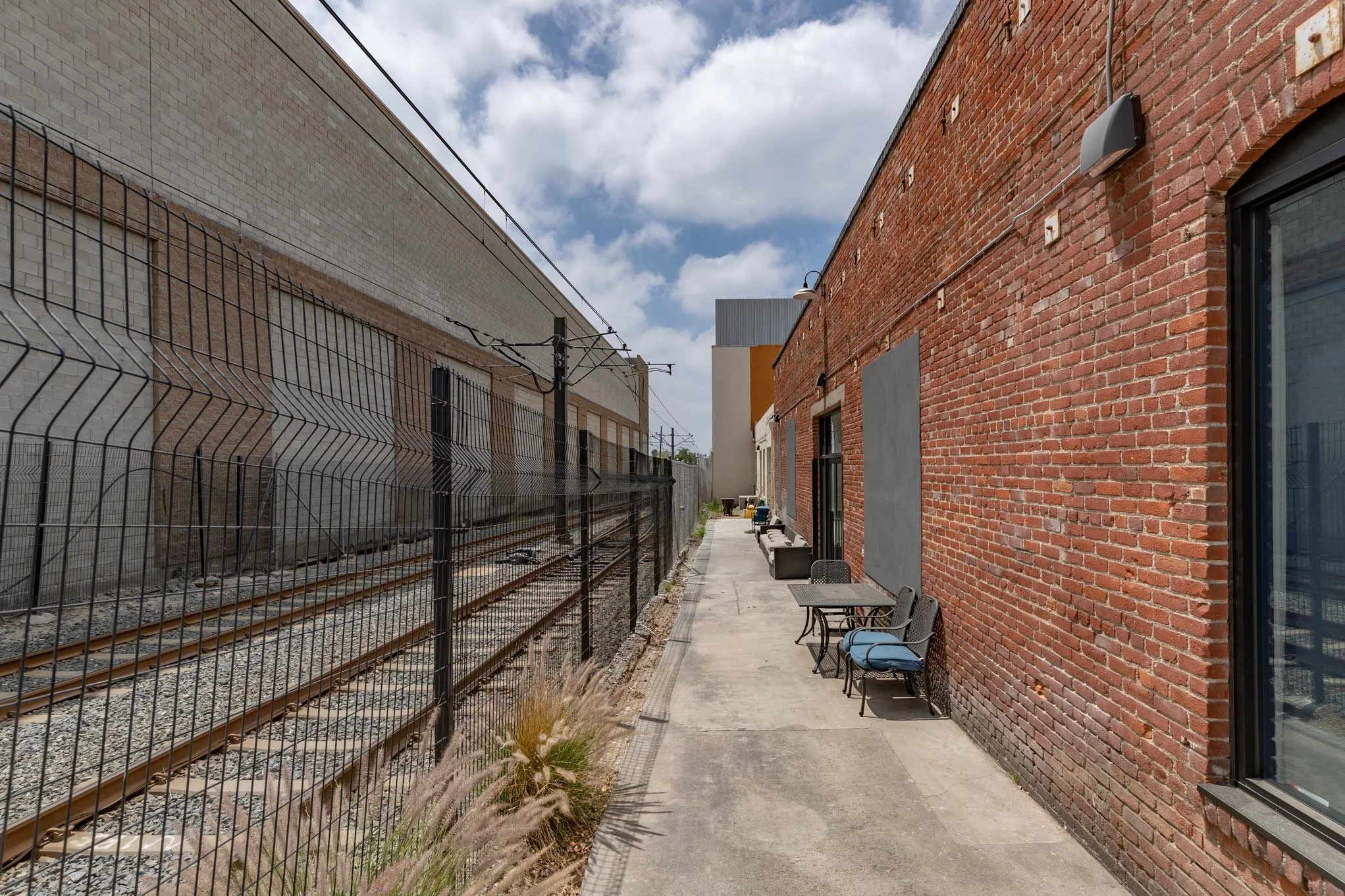 An alleyway between a brick building and a building on a fence-lined railway, with chairs and tables on the sidewalk.