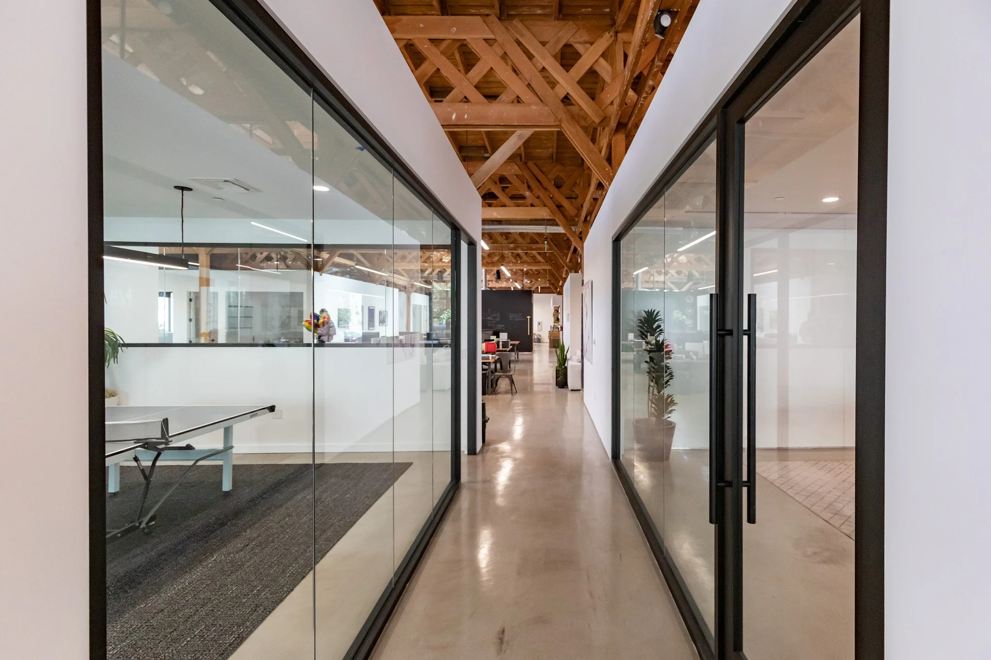 A modern office hallway with glass-walled rooms on both sides, wooden ceiling beams, and a polished concrete floor.