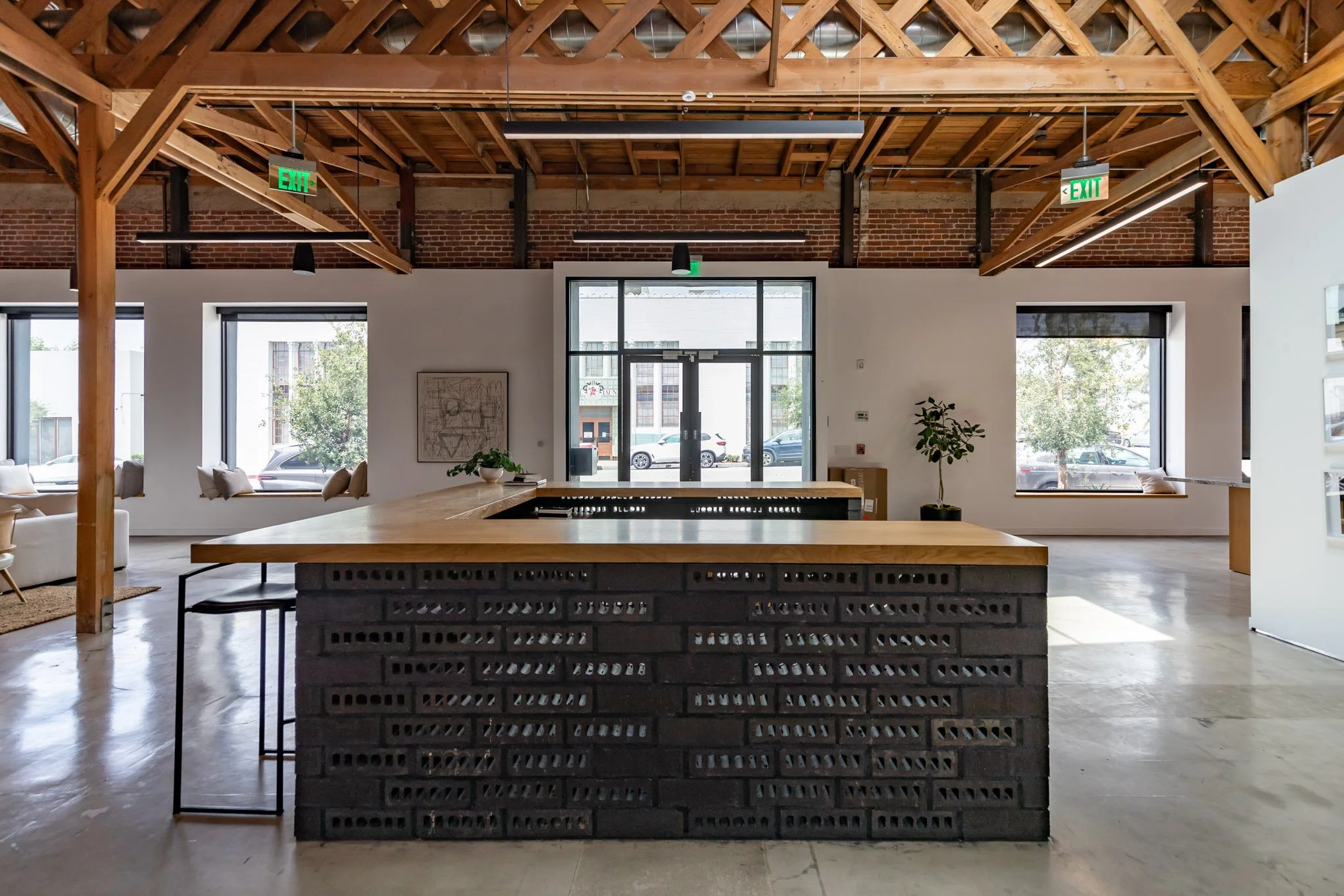 Open office space with a large kitchen island made of black bricks with a wooden countertop, large windows, and exposed wooden beams in the ceiling.