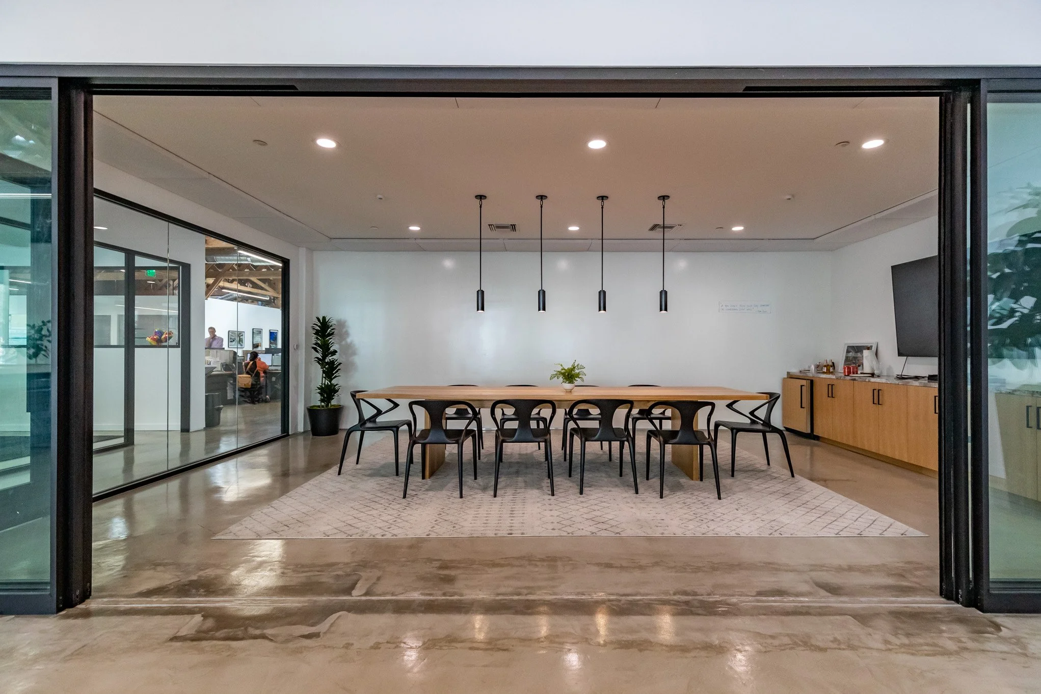 Empty modern conference room with a large wooden table, black chairs, a white wall with four hanging pendant lights, and a sideboard with a TV and items on top. There are plants and a patterned rug under the table.