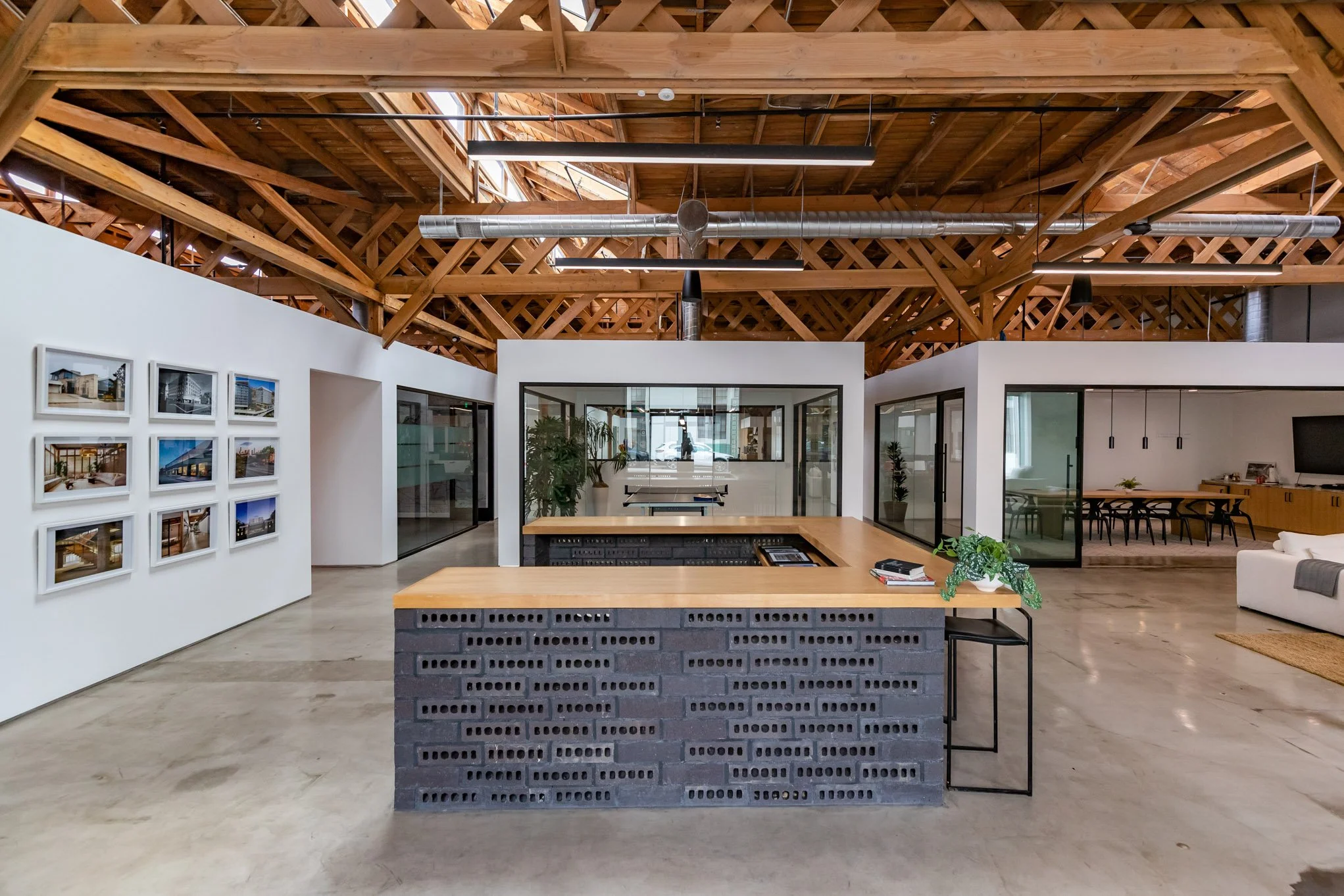 Modern open office space with a central brick and wood reception desk, picture frames on the white wall, glass-walled rooms, exposed wooden ceiling beams, and ductwork.