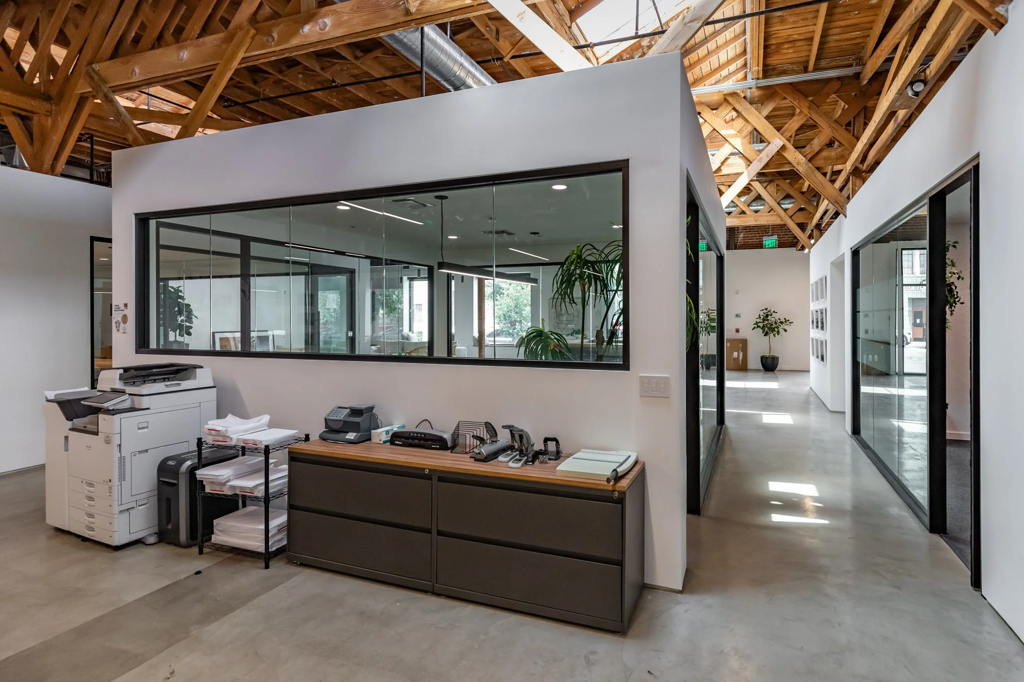 Modern office reception area with glass walls, wooden ceiling beams, and office equipment including printers and phones on a desk.