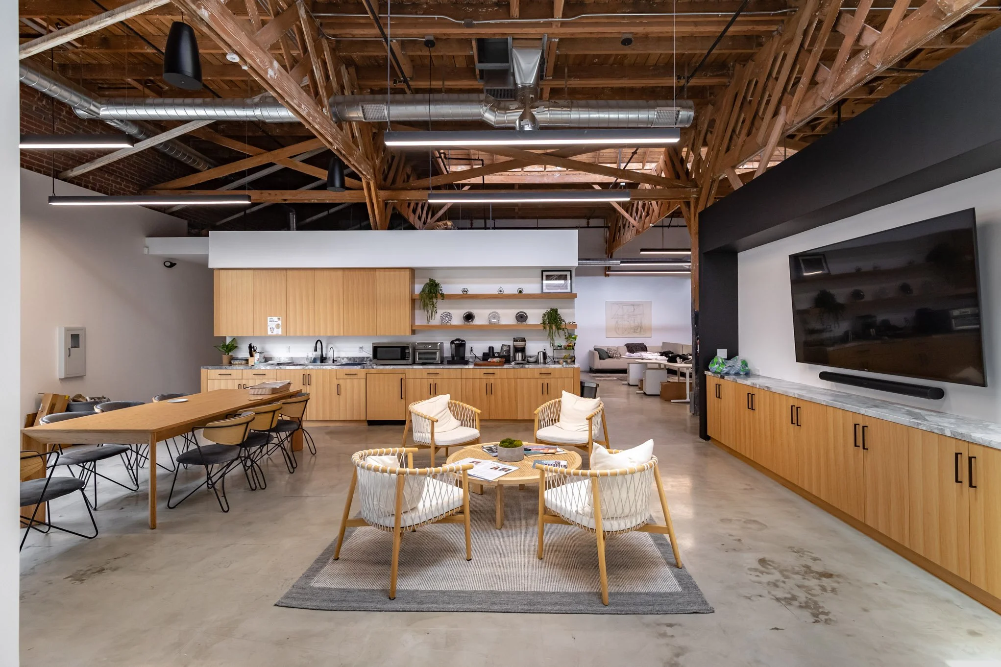 Open concept lounge with a cozy seating area, wooden cabinetry, a large flat-screen TV, and a kitchen with a wooden table, chairs, and modern appliances, featuring exposed wooden beams and ductwork on the ceiling.
