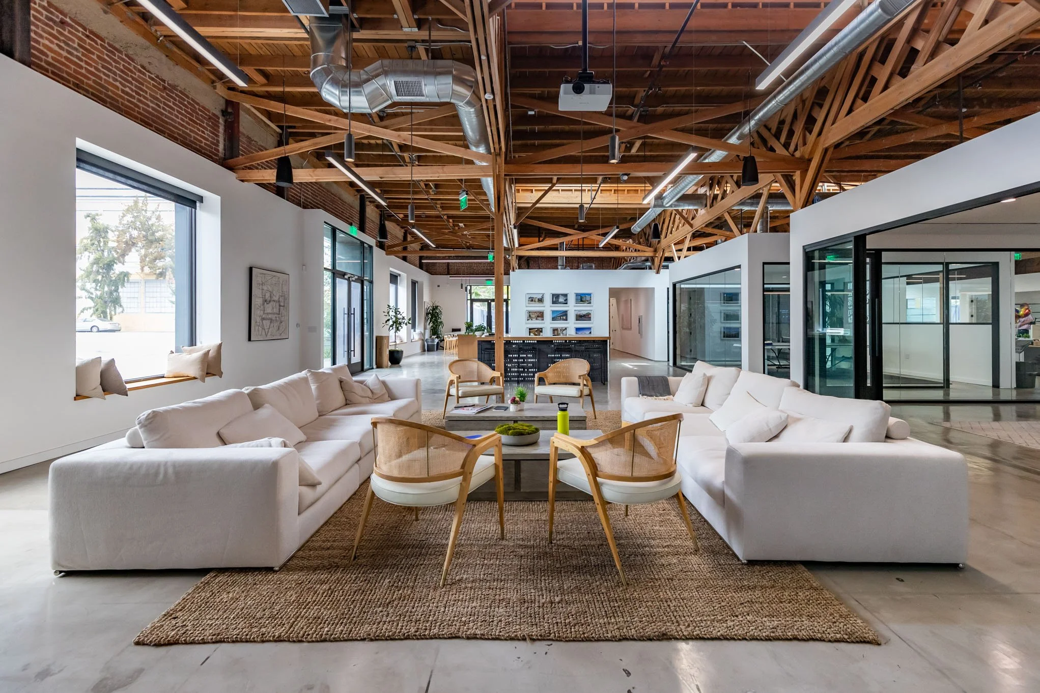 Modern office or lounge area with white sofas, wooden chairs, and a central coffee table on a woven rug, with large windows, exposed wooden beams, and glass-enclosed offices visible in the background.