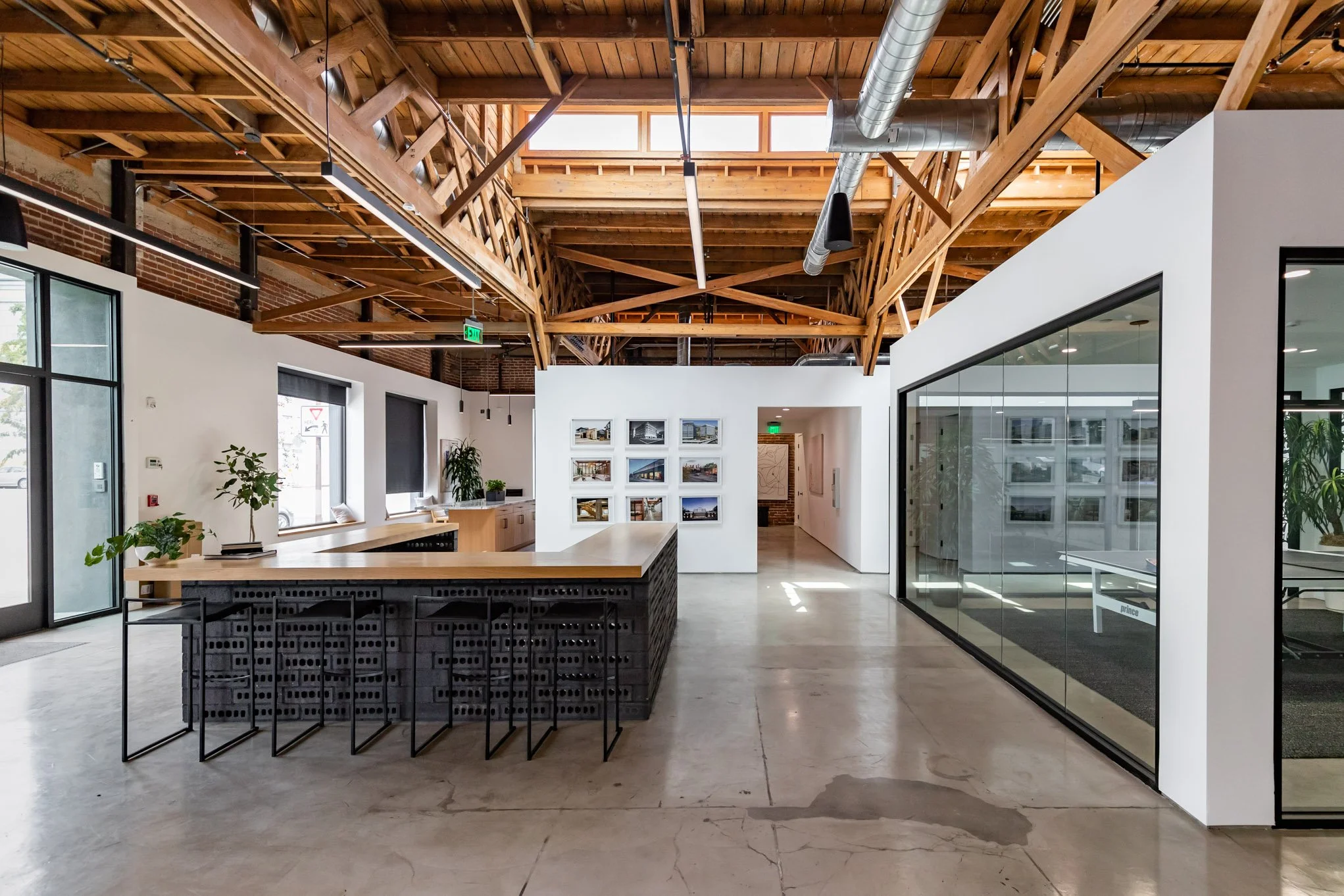 Modern industrial-style office lobby with exposed wooden beams, concrete floor, glass walls, plants, and a reception desk.