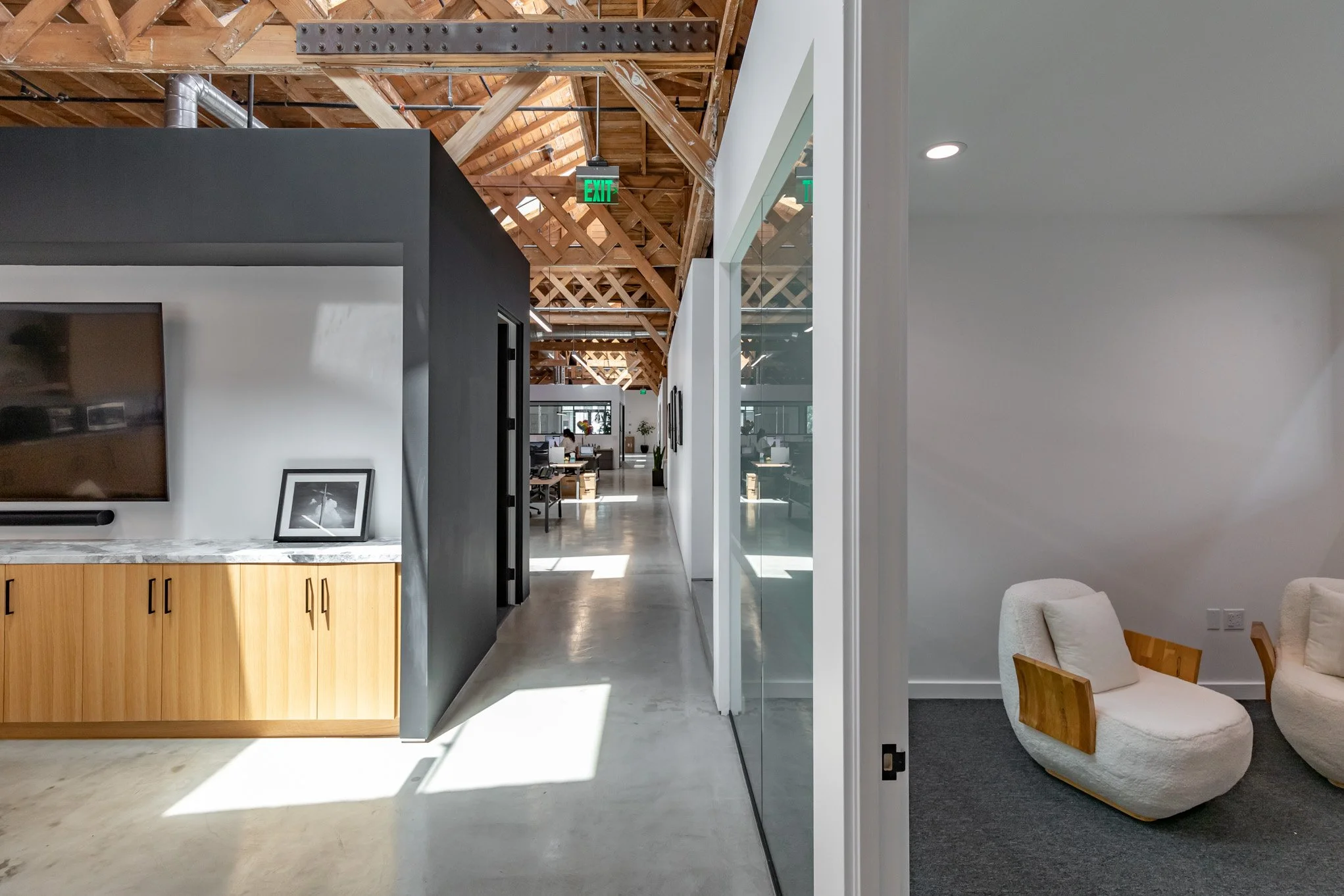 Interior of a modern office with white walls, wooden furniture, and a visible ceiling with wooden beams and ducts, partially separated by glass walls, with a seating area on the right that includes two plush chairs with wooden armrests.