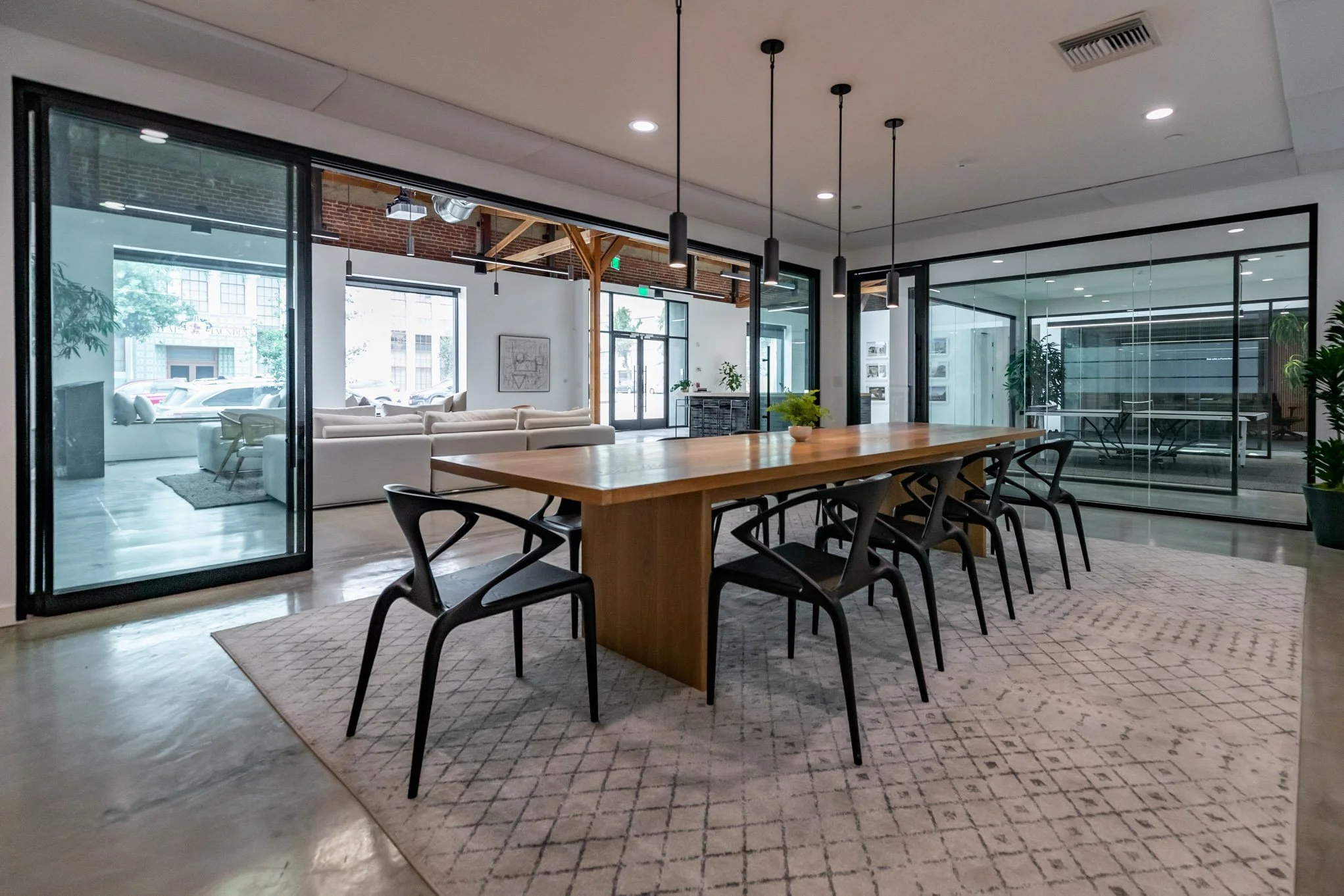 Modern conference room with a long wooden table, black chairs, and large glass walls showing a lounge area with white sofas and large windows.