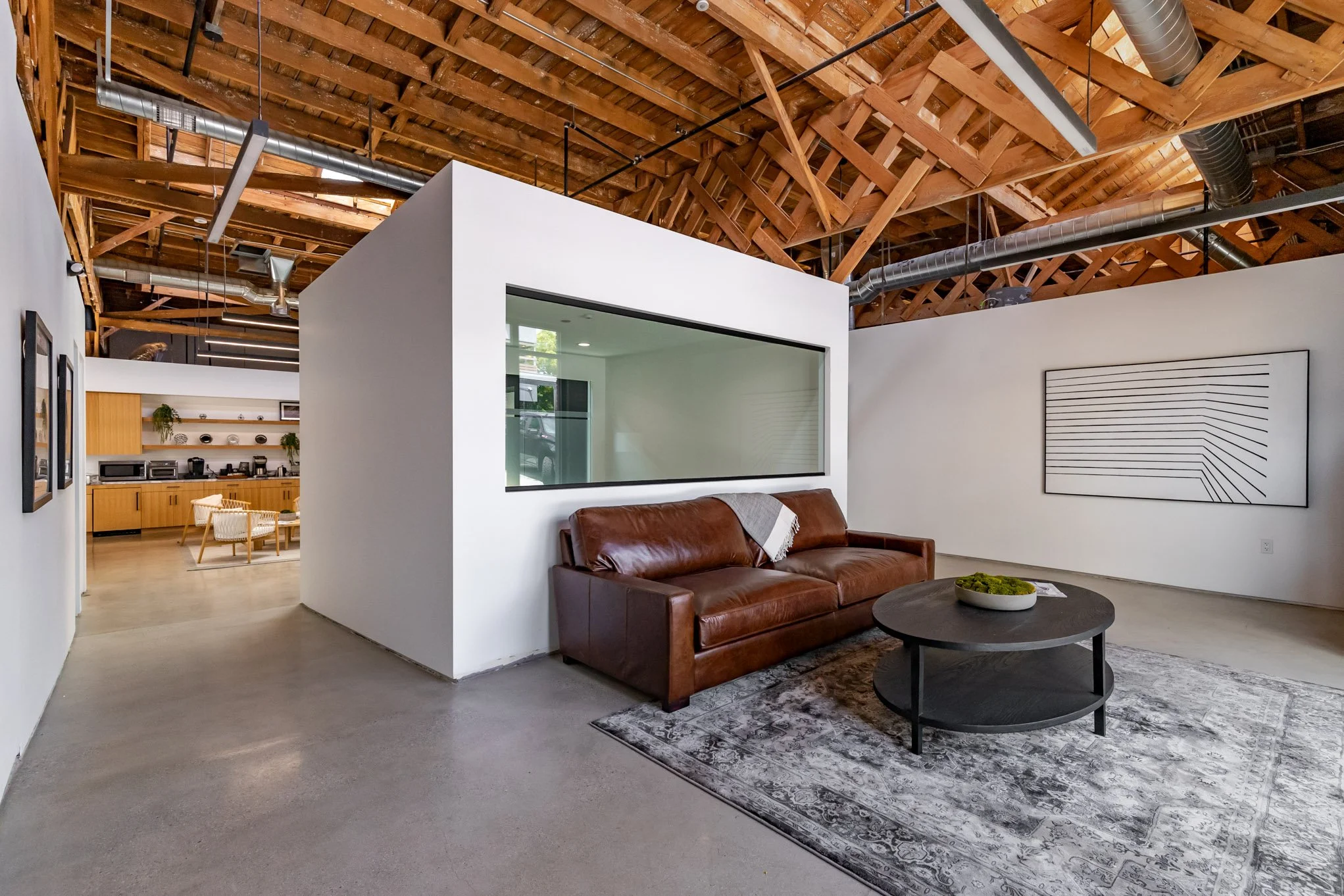 Modern lounge area with a brown leather sofa, a black oval coffee table on a patterned rug, and white walls with artwork, featuring industrial-style exposed wooden ceiling beams.