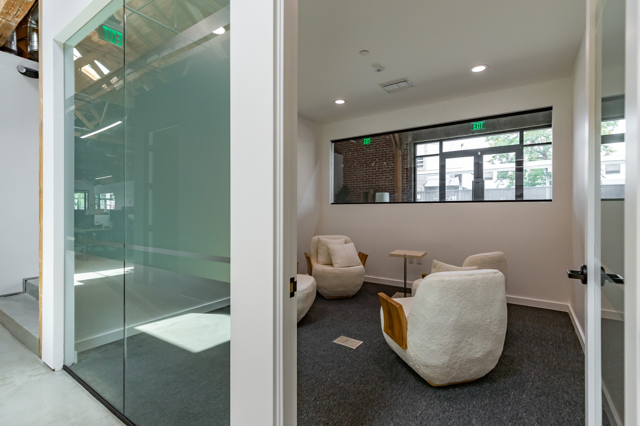 Interior view of a modern office lounge area with white plush chairs, a small table, and large windows showing a brick wall and outdoor scene.