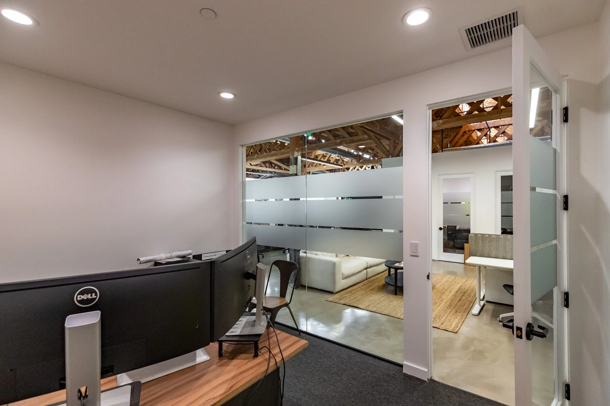 Office interior with a curved desk and a dual-monitor setup in the foreground, and a glass-walled meeting room with white couches and a wooden coffee table in the background.