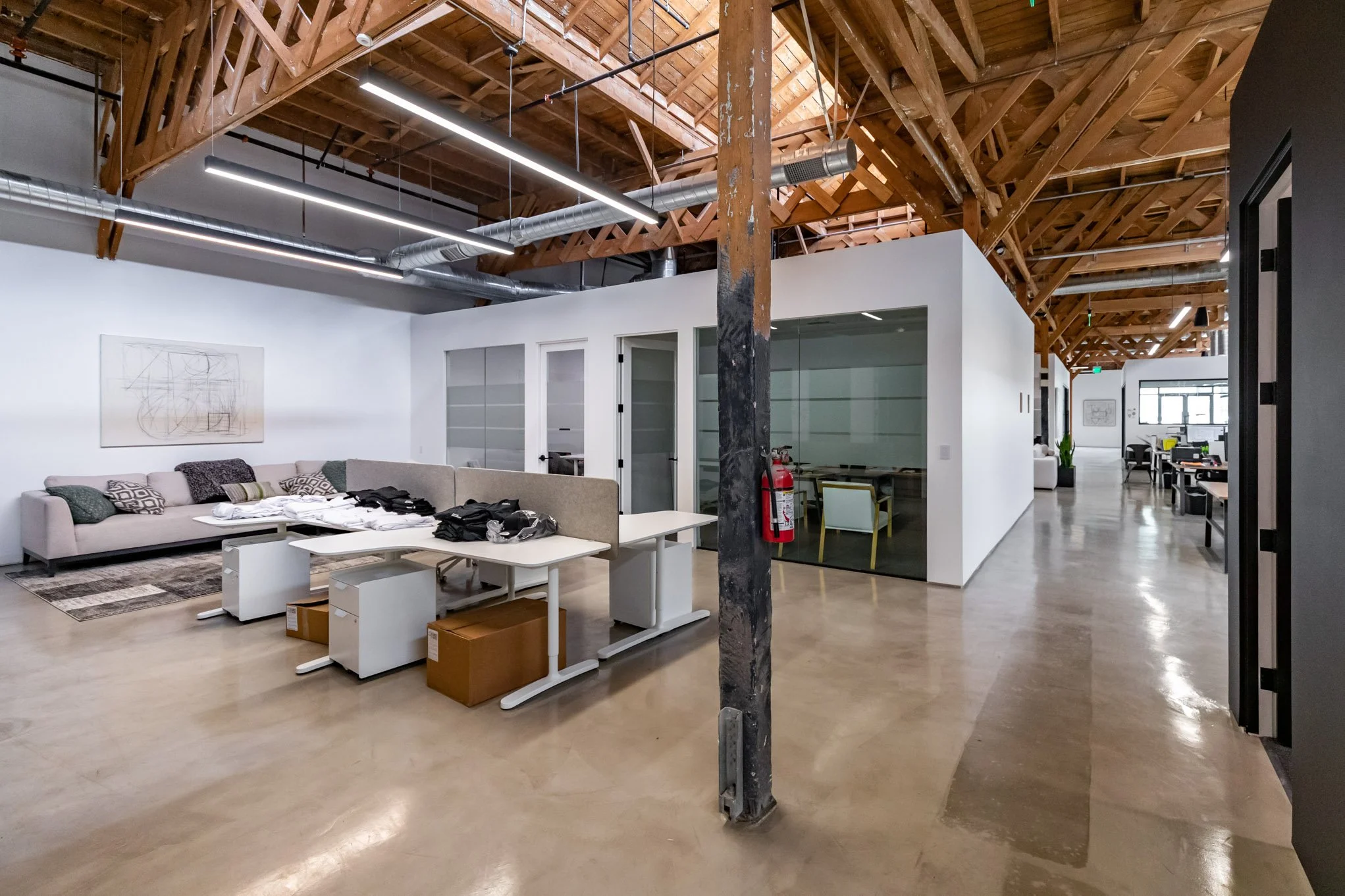 Open office space with wooden ceiling, white walls, and polished concrete floors. There is a large gray sofa with pillows and a small abstract artwork on the wall. In front are several white desks and chairs, some with boxes underneath. To the right 