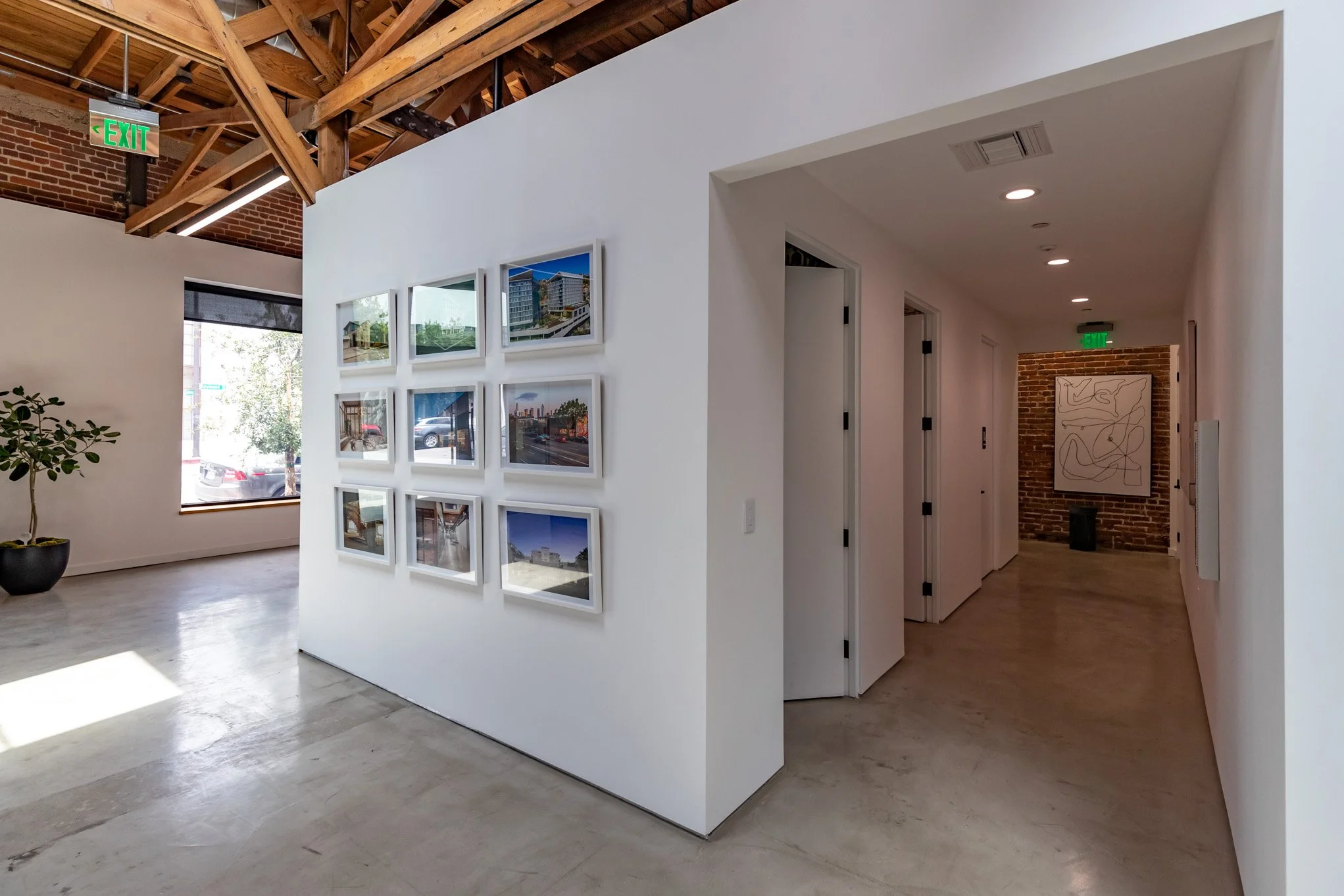Interior of an art gallery with a white wall displaying nine framed photographs, a hallway with closed doors, an exposed brick wall at the end with abstract art, and a potted plant near a large window.