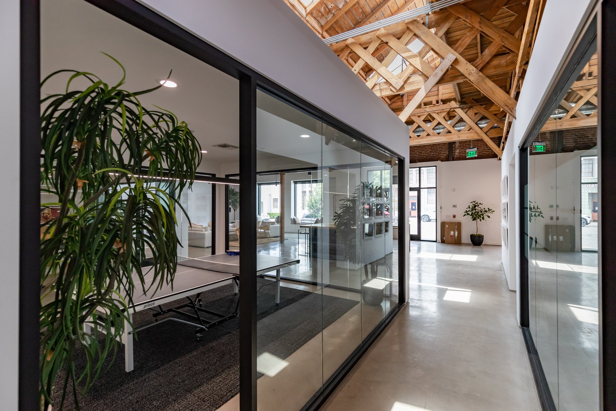Modern office space with glass partition walls, wooden ceiling beams, potted plants, and natural lighting.