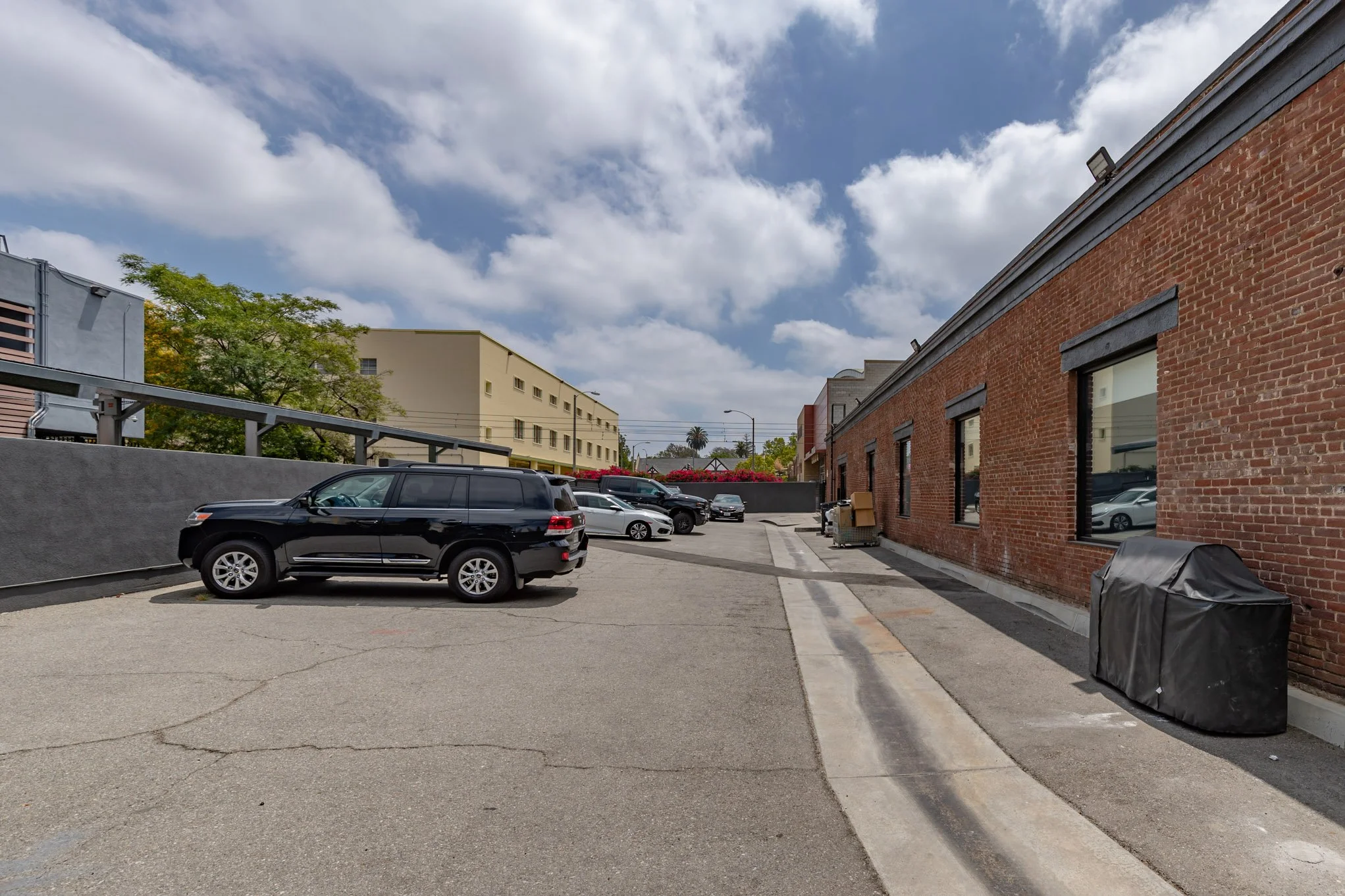 A parking lot next to a brick building with several parked cars and a sidewalk, under a partly cloudy sky.