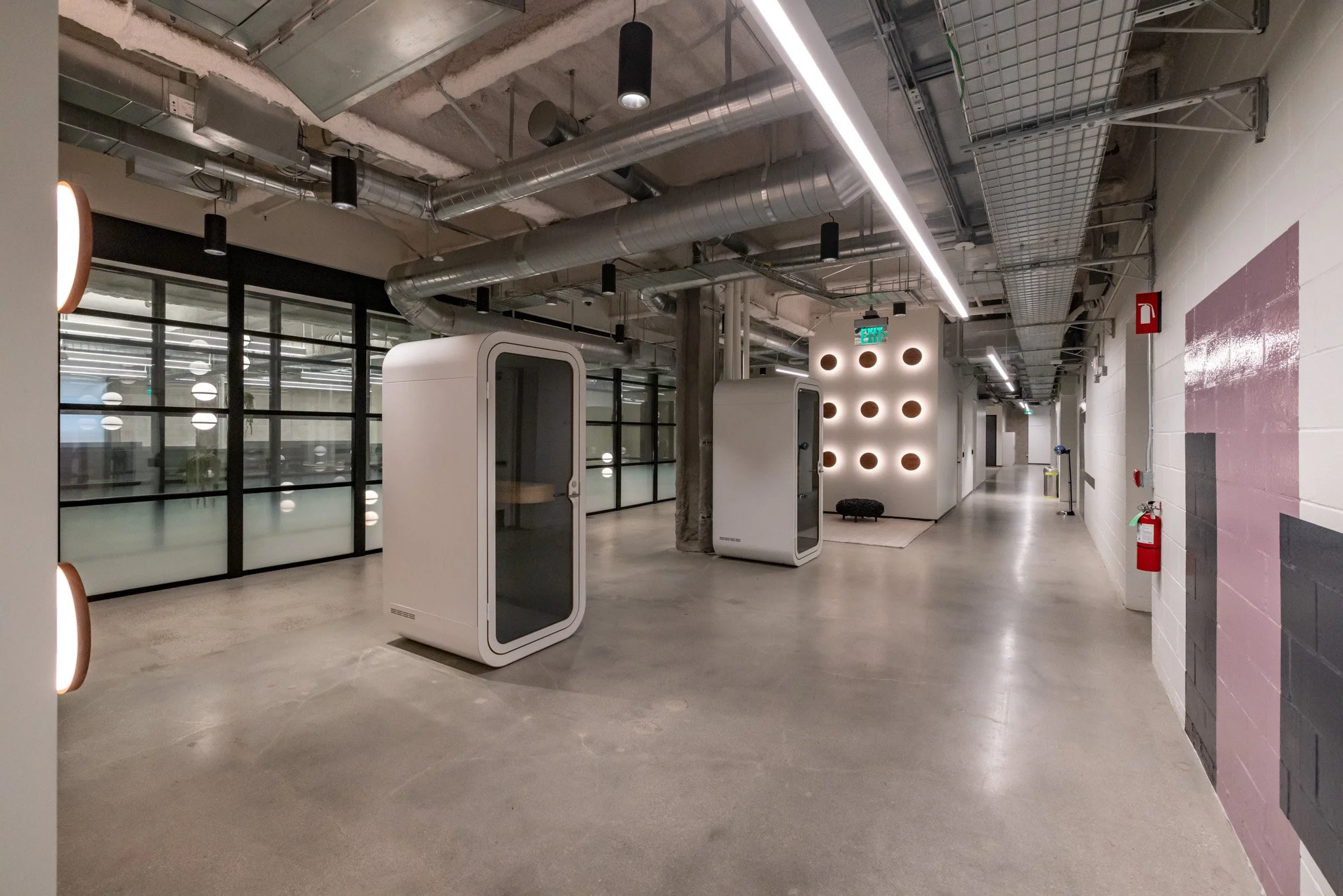 Modern office corridor with exposed ceiling pipes, glass partitions, and isolated phone booths. Decor includes round wall lights and a small seating area.