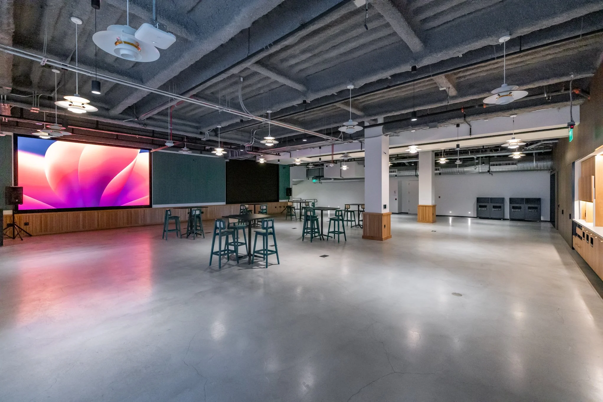 Empty modern event space with a large digital screen, high ceilings with exposed pipes and fans, multiple tables with chairs, and a polished concrete floor.