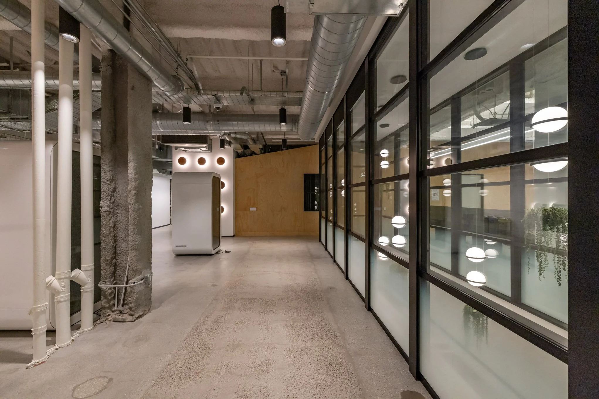 Modern interior space with exposed industrial ceiling, white and black pipes, a large glass partition with black framing, decorative lighting fixtures, and a wooden wall.