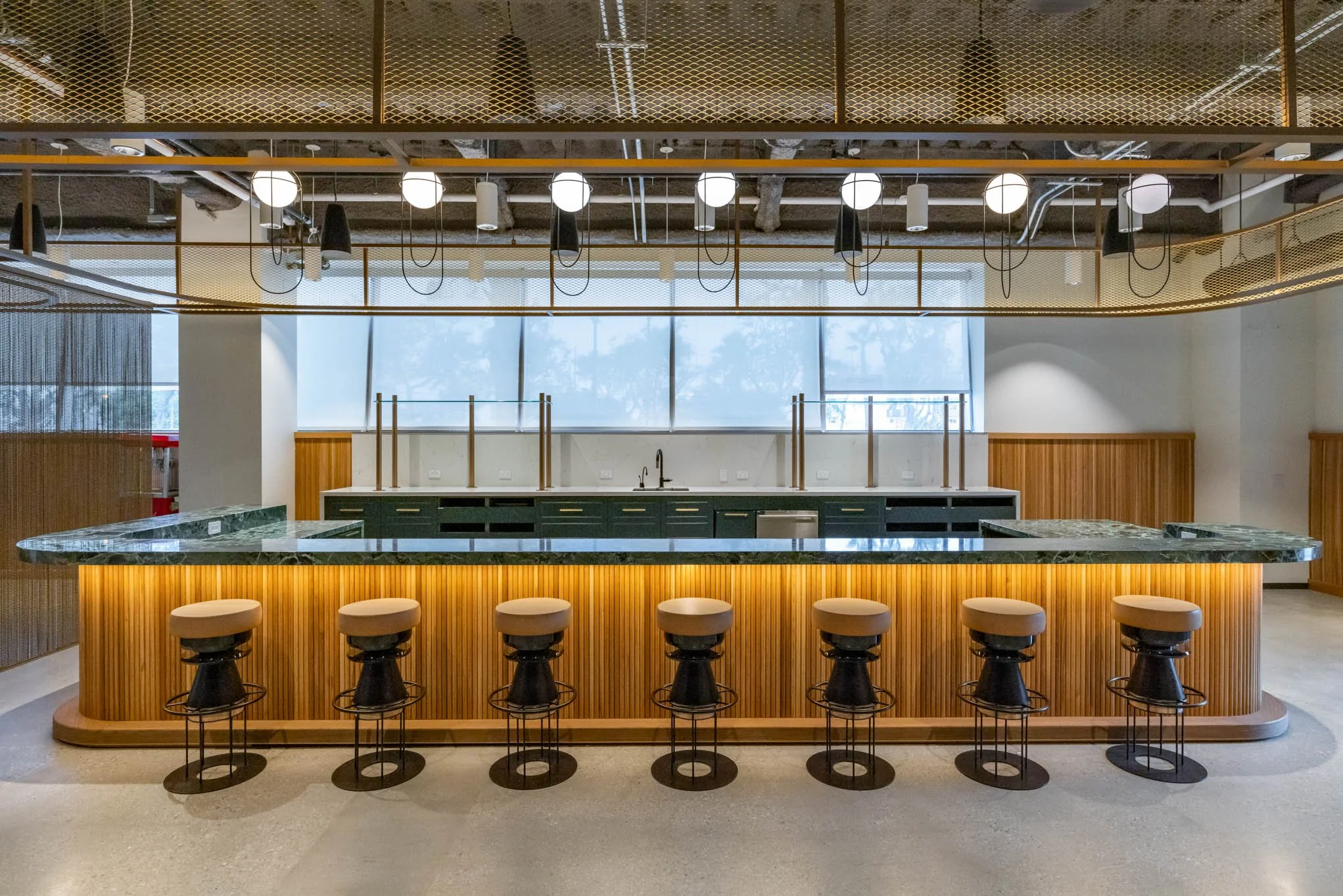 Modern, empty bar with a marble countertop, six high stools, green cabinets, and large windows in the background.