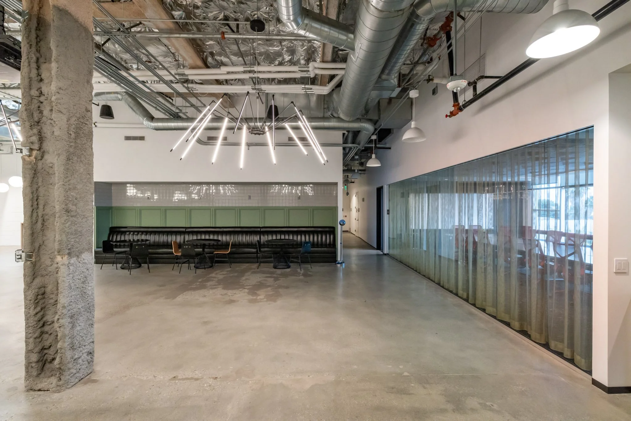 Modern, industrial-style interior space with concrete flooring, exposed ductwork, a curved black leather bench, assorted chairs, a glass wall with sheer curtains, and a contemporary light fixture hanging from the ceiling.