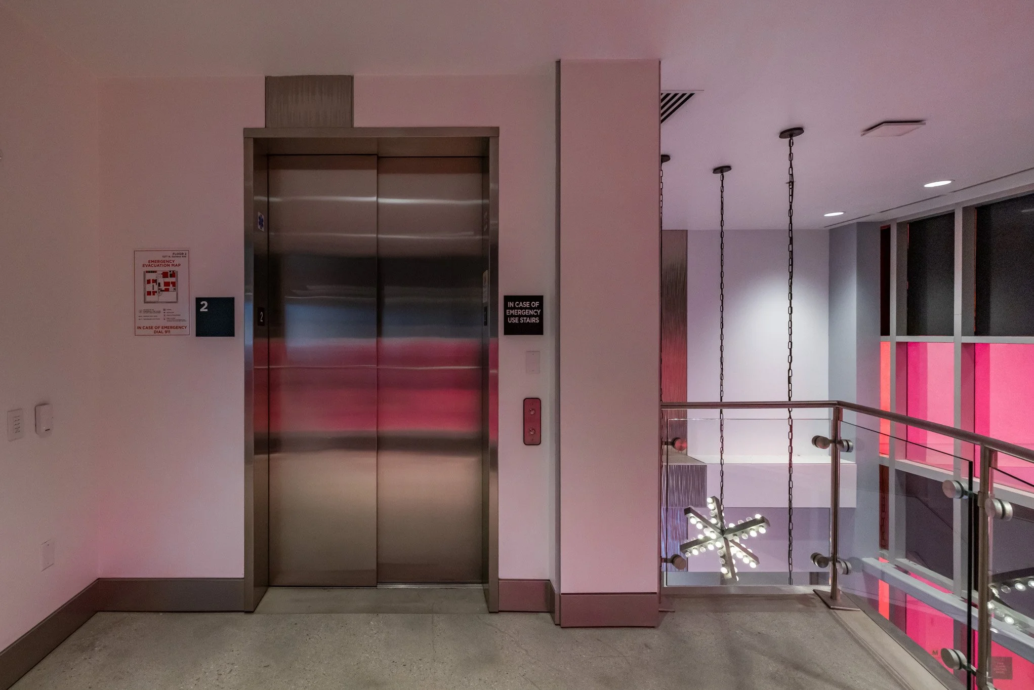 Elevator door next to a wall with emergency instructions and a sign, located in a modern building with pink lighting accents and a staircase with glass railing.