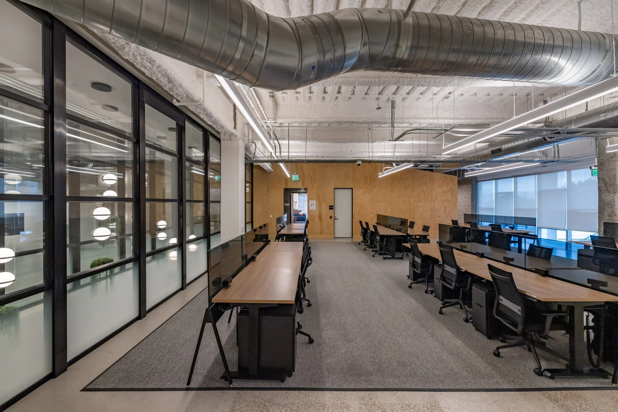 Modern office conference room with long desks, black chairs, glass partitions, and industrial ceiling ducts.