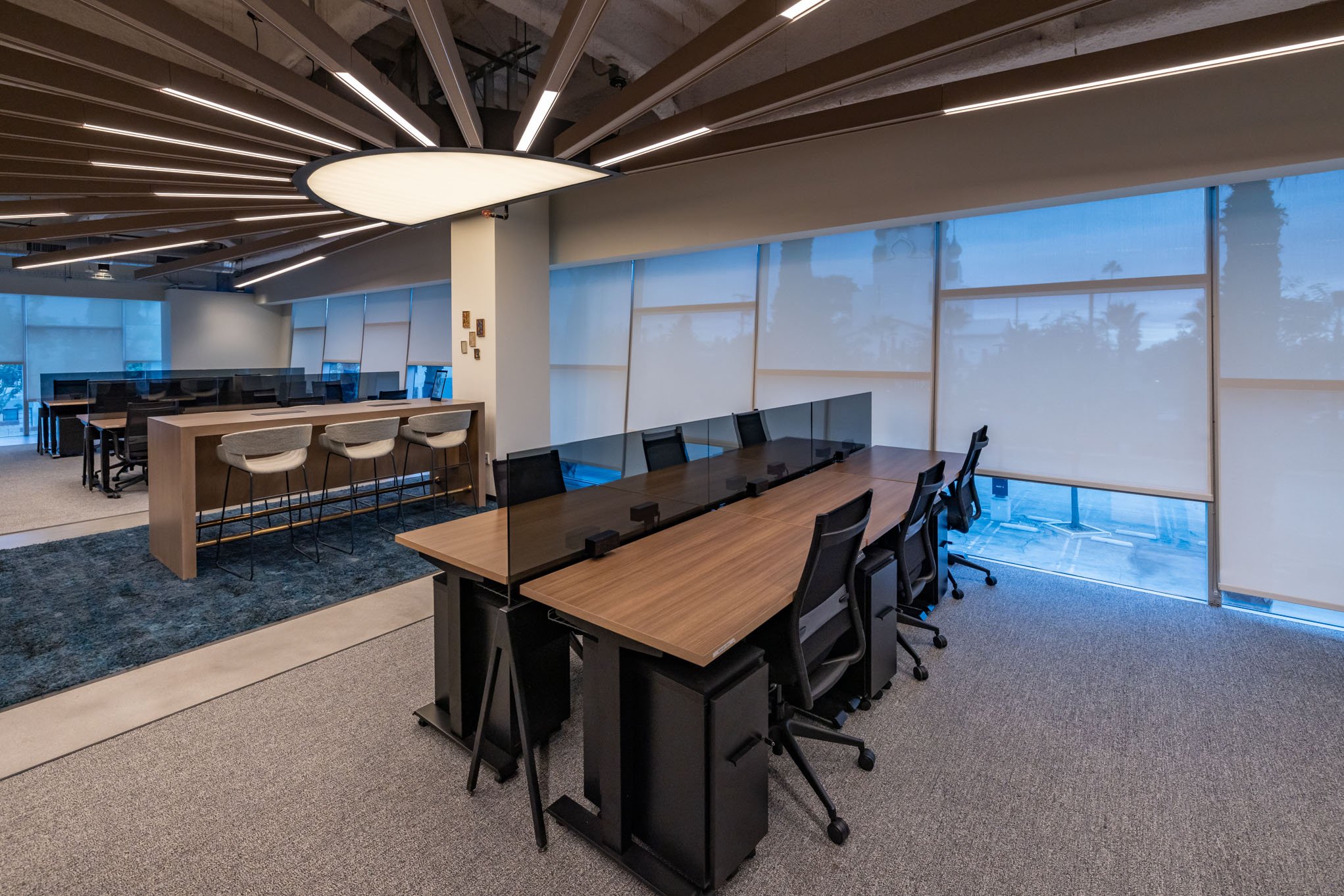 Modern office with a large rectangular table, black chairs, a glass partition, and a row of workstations in the background, illuminated by a contemporary ceiling light.