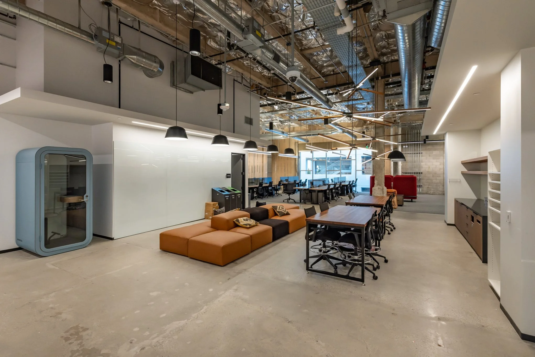 Modern office space with open layout, featuring a lounge area with orange and black seating, a long wooden table with black chairs, multiple workstations with monitors, exposed ceiling pipes and ducts, and a large window letting in natural light.