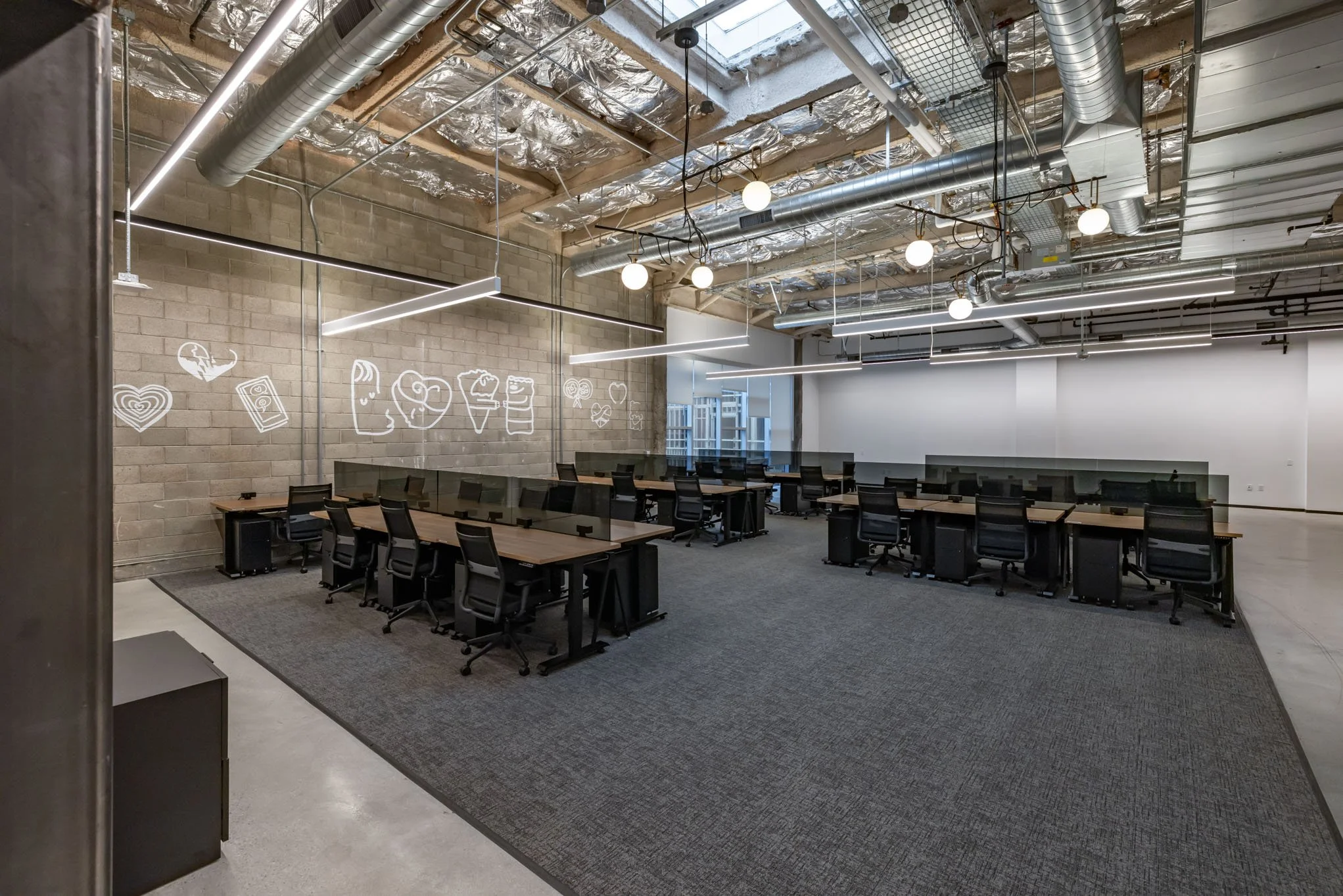Modern empty office with rows of black office chairs and desks separated by transparent dividers, exposed ceiling with ductwork, decorative chalk drawings of hearts and ice cream on concrete wall, and a large window.