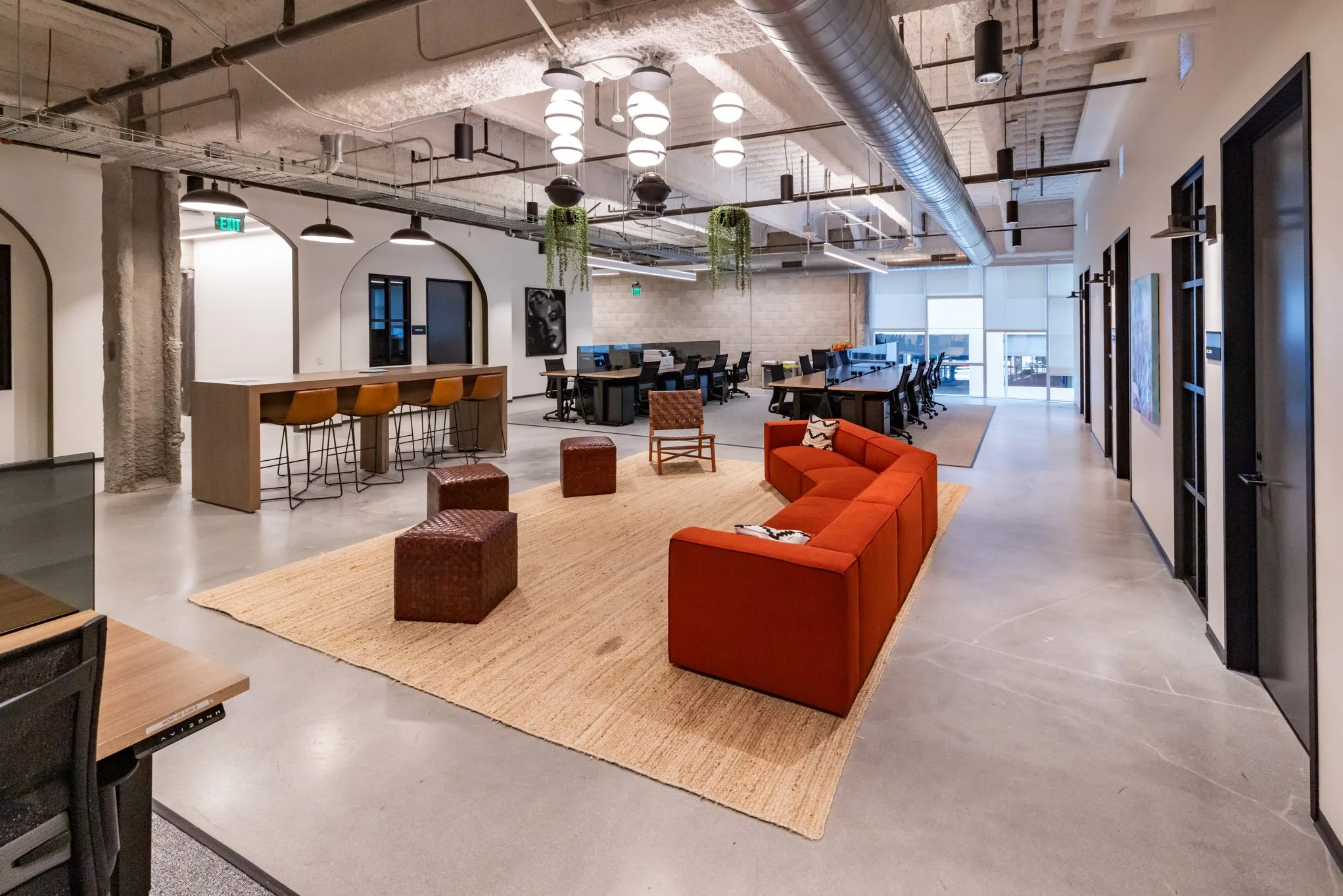Open-plan modern office with a seating area that has an orange sectional sofa, two small woven ottomans, and a wooden chair on a large beige rug. In the background, there are workstations with computers and black chairs, and an area with a high count