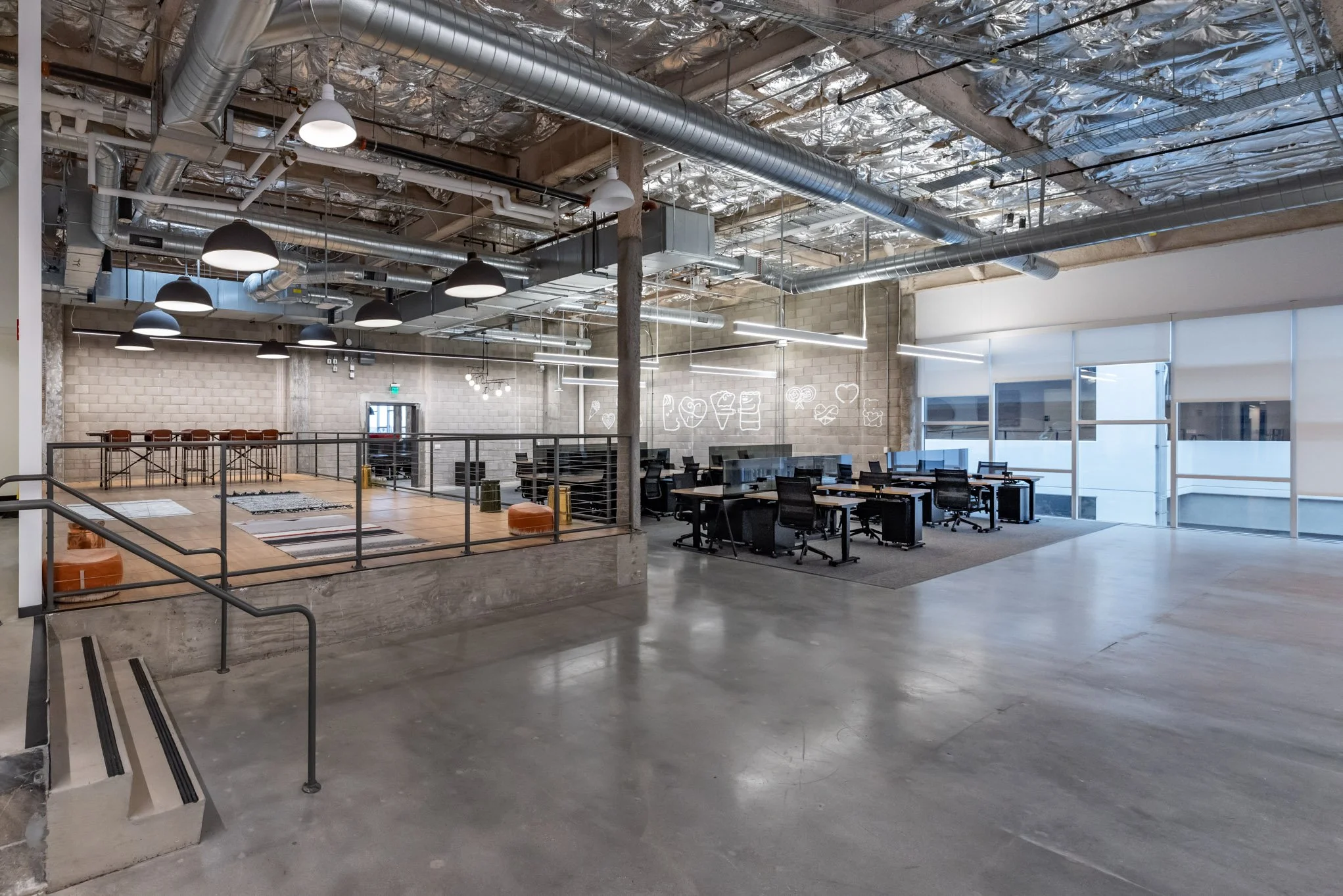 Open-concept coworking space with a raised rug area, multiple desks with chairs, large windows, and modern industrial ceiling with exposed ductwork and lighting.