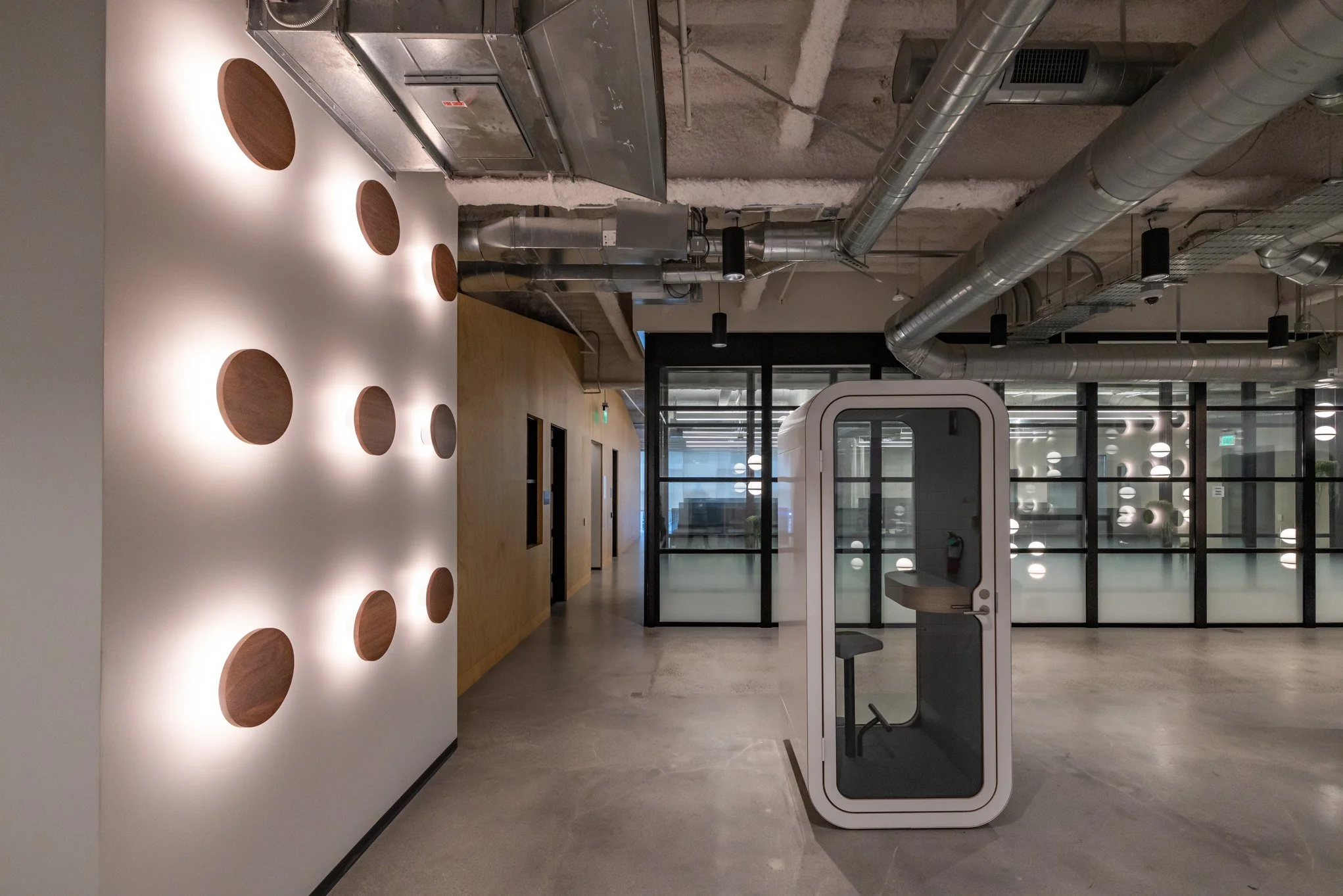 Modern office interior with a glass-walled meeting pod, wooden wall with circular cutouts, and exposed ceiling pipes.