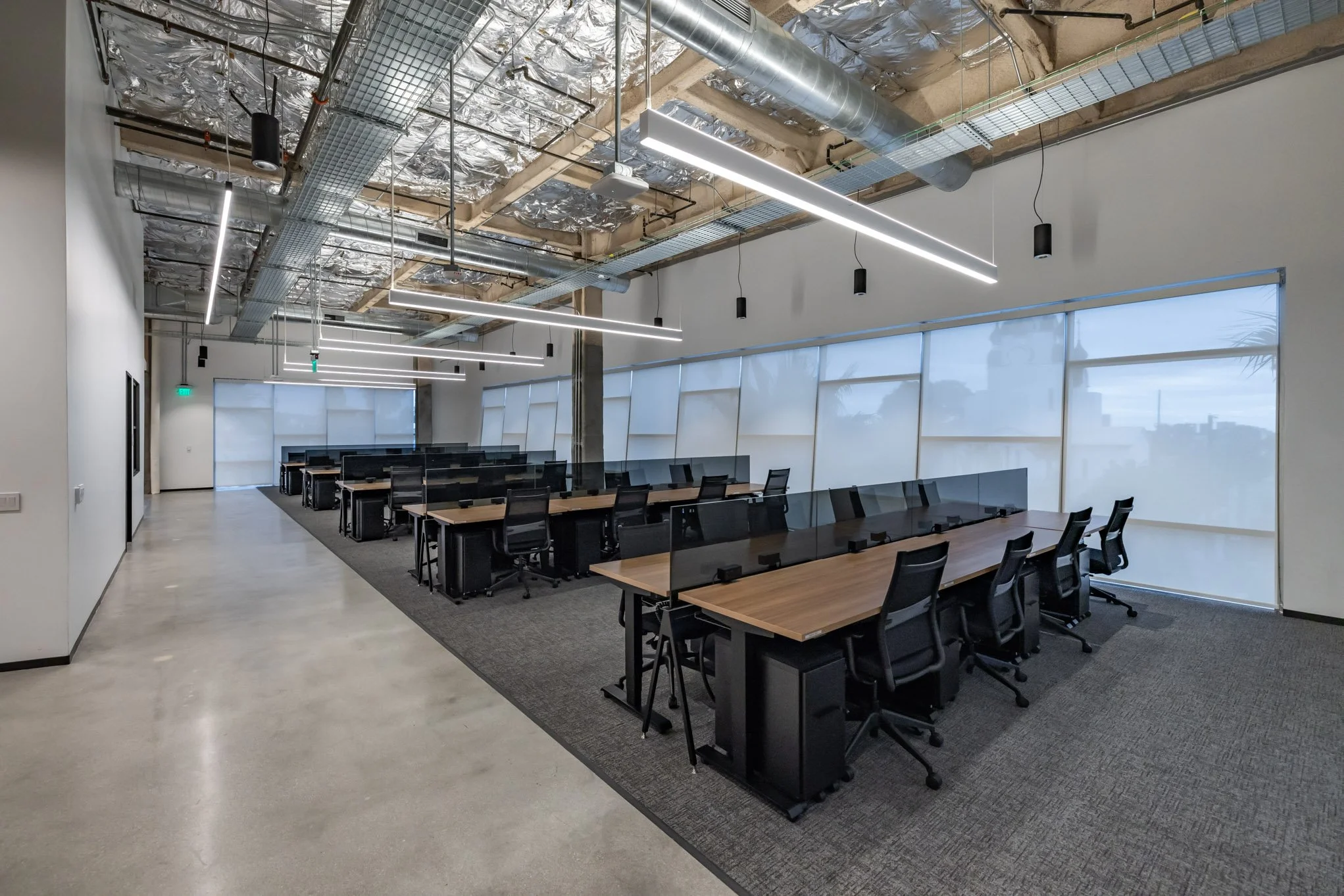 Empty modern coworking space with desks, chairs, and glass dividers, industrial ceiling with exposed pipes and ducts, large windows with blinds showing an overcast sky.