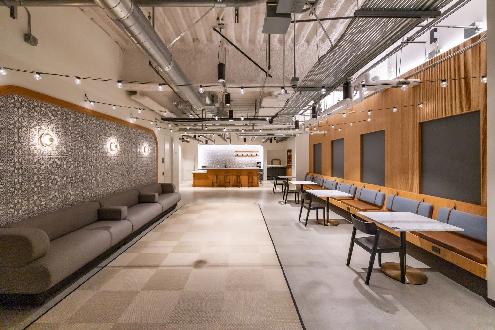 A modern, industrial-style lounge with a long gray sofa on the left, a wooden wall with blue cushioned benches and tables on the right, a patterned wall with lights, and a bar with stools at the far end. Exposed ductwork and piping are visible on the