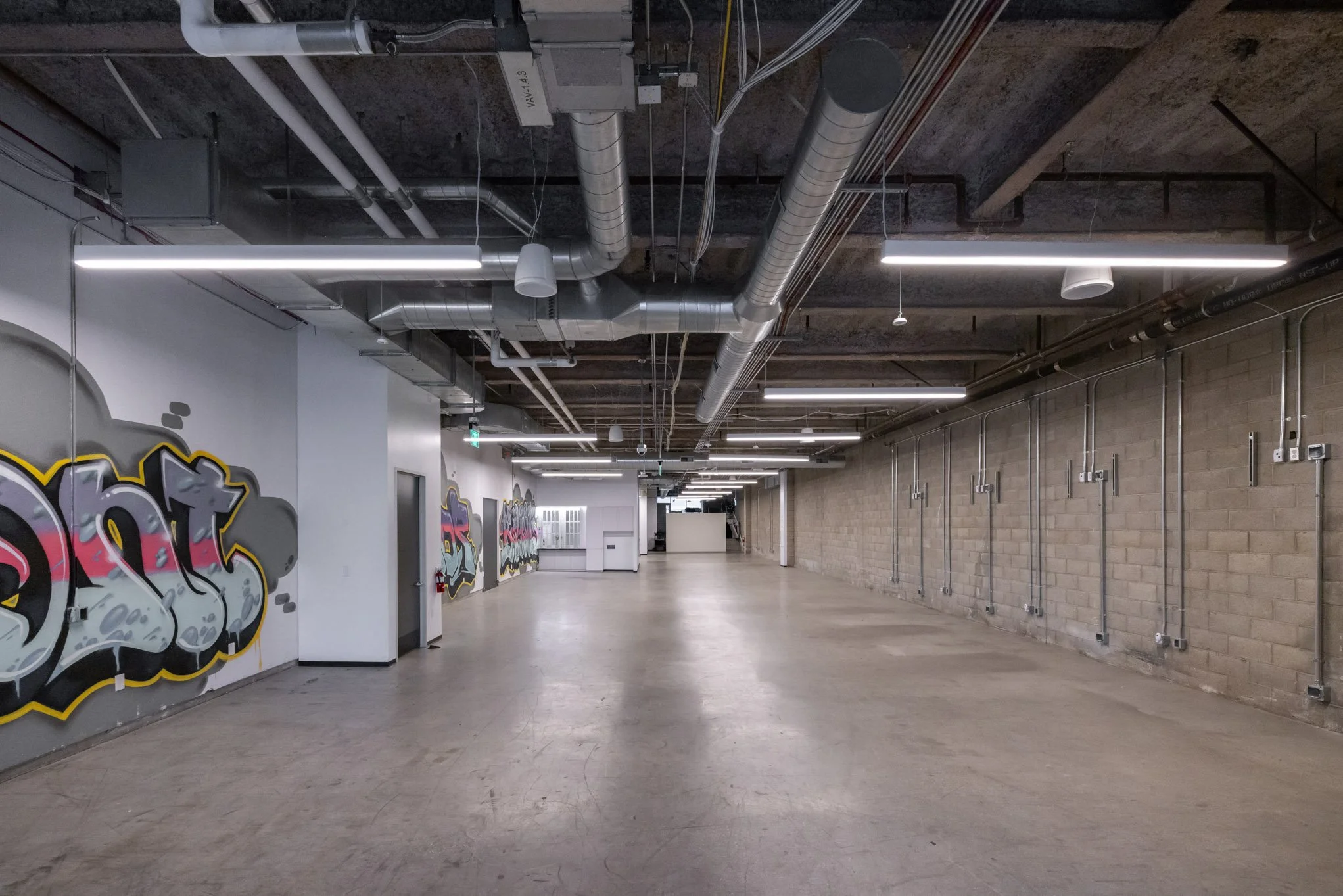 Empty industrial-style parking garage with graffiti on the wall, exposed pipes, and linear ceiling lighting.