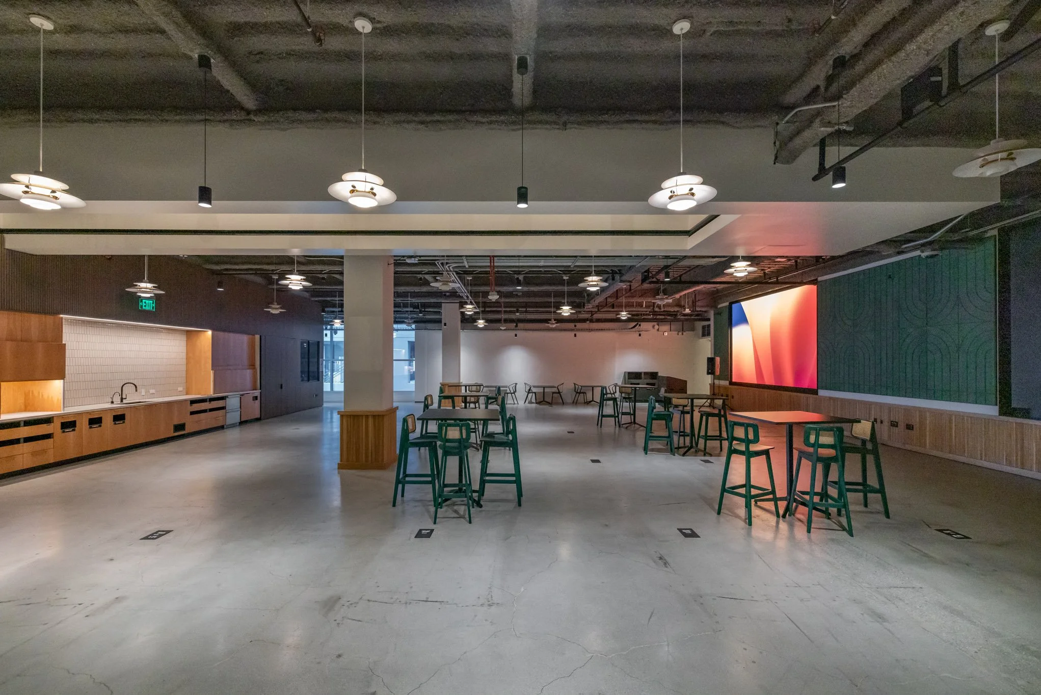 Empty modern cafe or lounge area with high tables, green stools, wooden accents, a large screen, and ceiling lights.