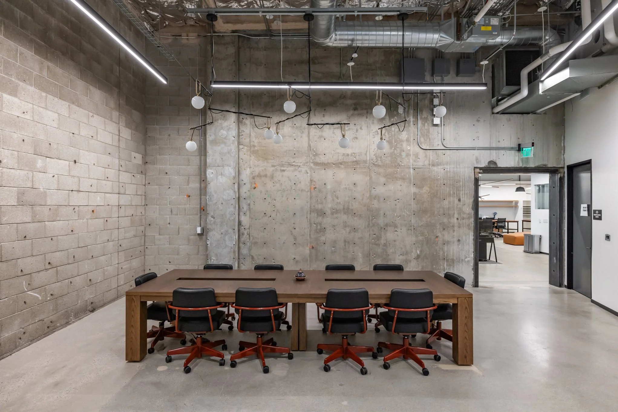 Conference room with wooden table surrounded by ten black office chairs with orange bases in an industrial-style space with exposed concrete walls and ceiling, illuminated by linear LED lighting and decorative hanging light bulbs.