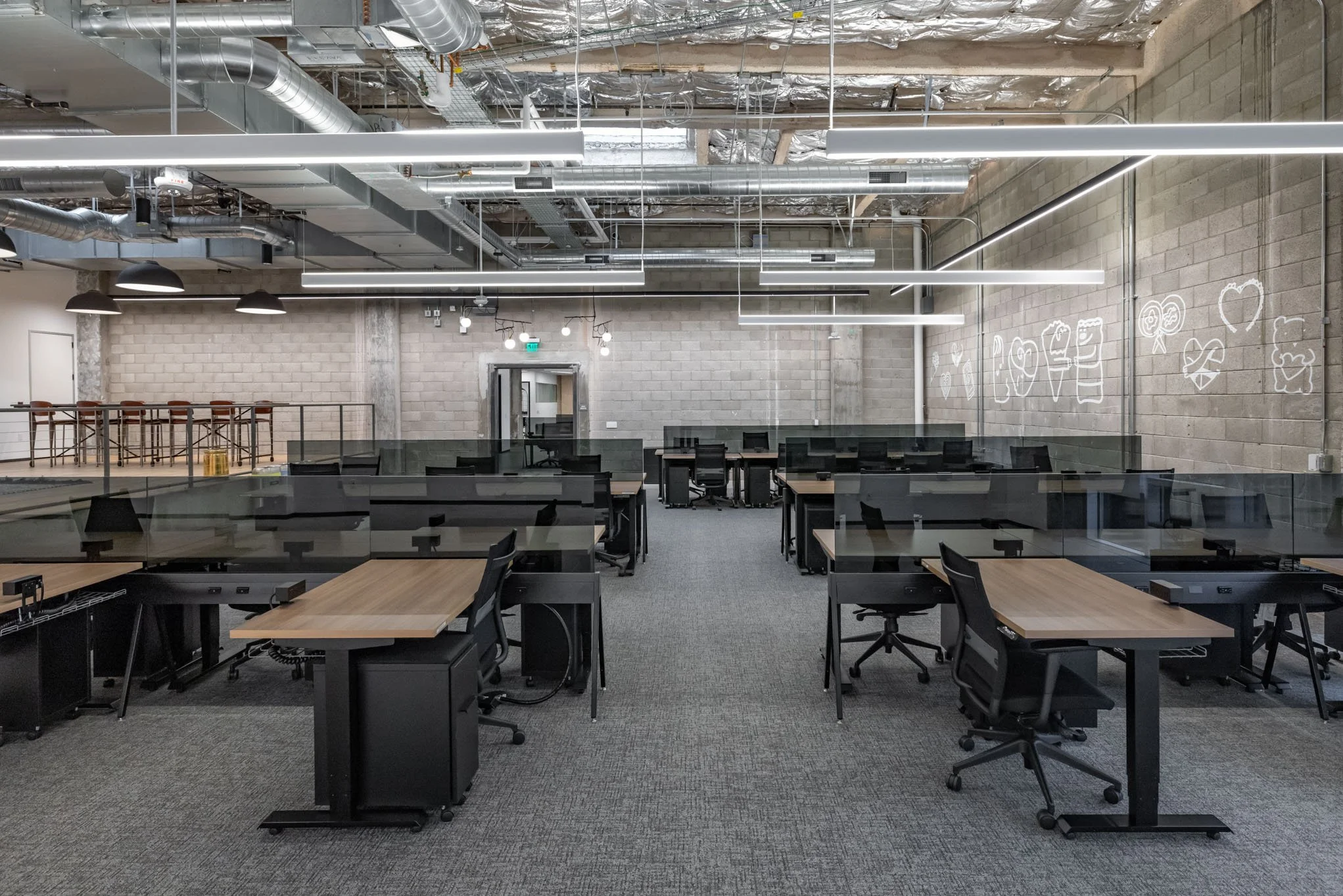 Empty modern office workspace with multiple desks and chairs, glass partitions, exposed ceiling pipes, wall chalk art, and industrial decor.