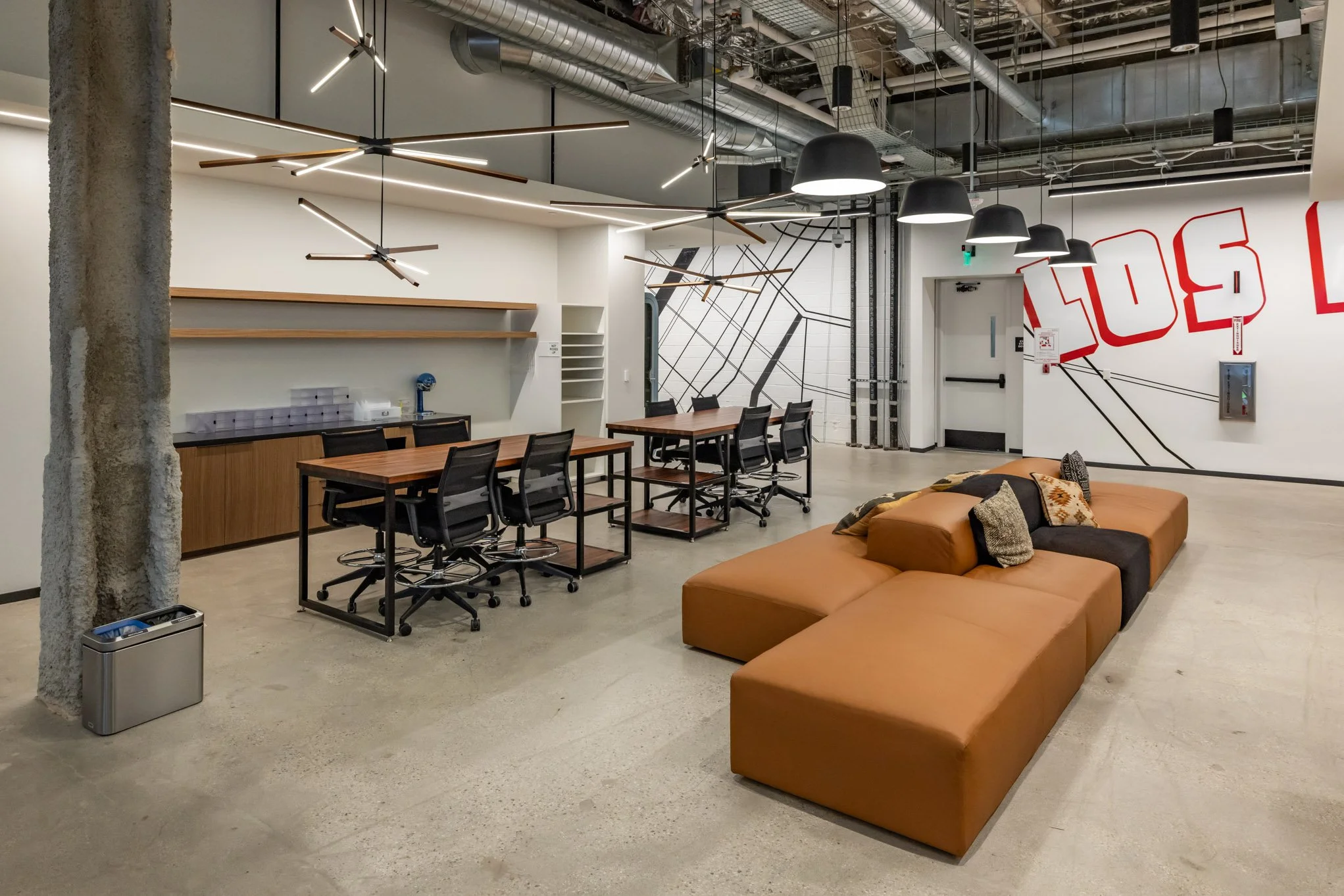 Modern office lounge area with a brown sectional sofa, black chairs at tables, contemporary lighting fixtures, exposed ductwork, a wall with red and black geometric mural, and a concrete floor.