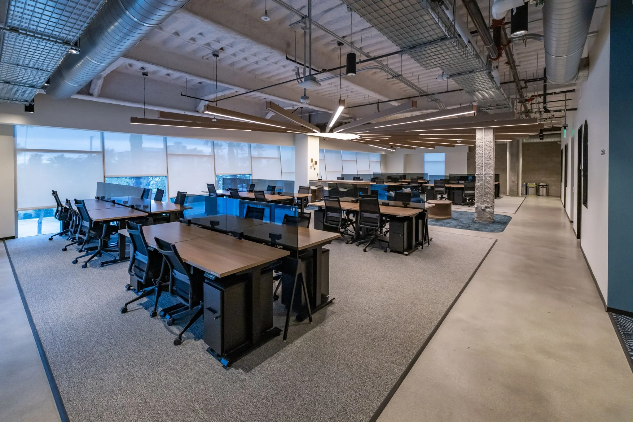 Modern open-plan office space with multiple desks, black office chairs, large windows with shades, and ceiling exposed ductwork and lighting fixtures.