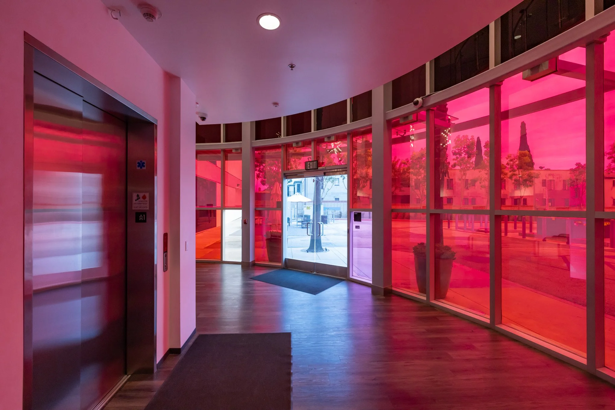 Interior of a building with glass walls tinted pink, showing an elevator, doors, and an outdoor patio with trees and umbrellas.