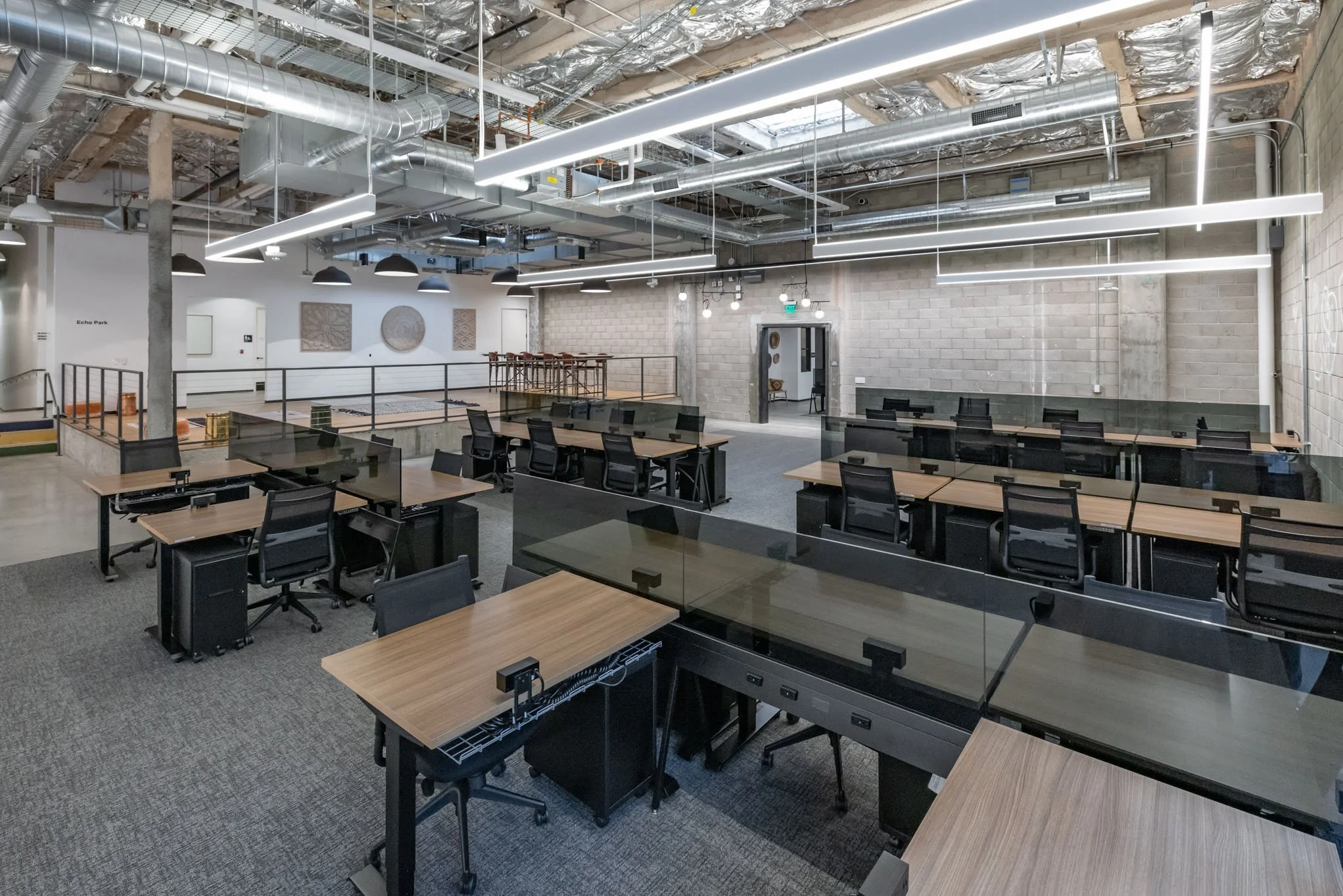 An open-concept modern office space featuring multiple rows of desks with black chairs, glass dividers, and exposed industrial ceiling elements with large ducts and lighting fixtures.