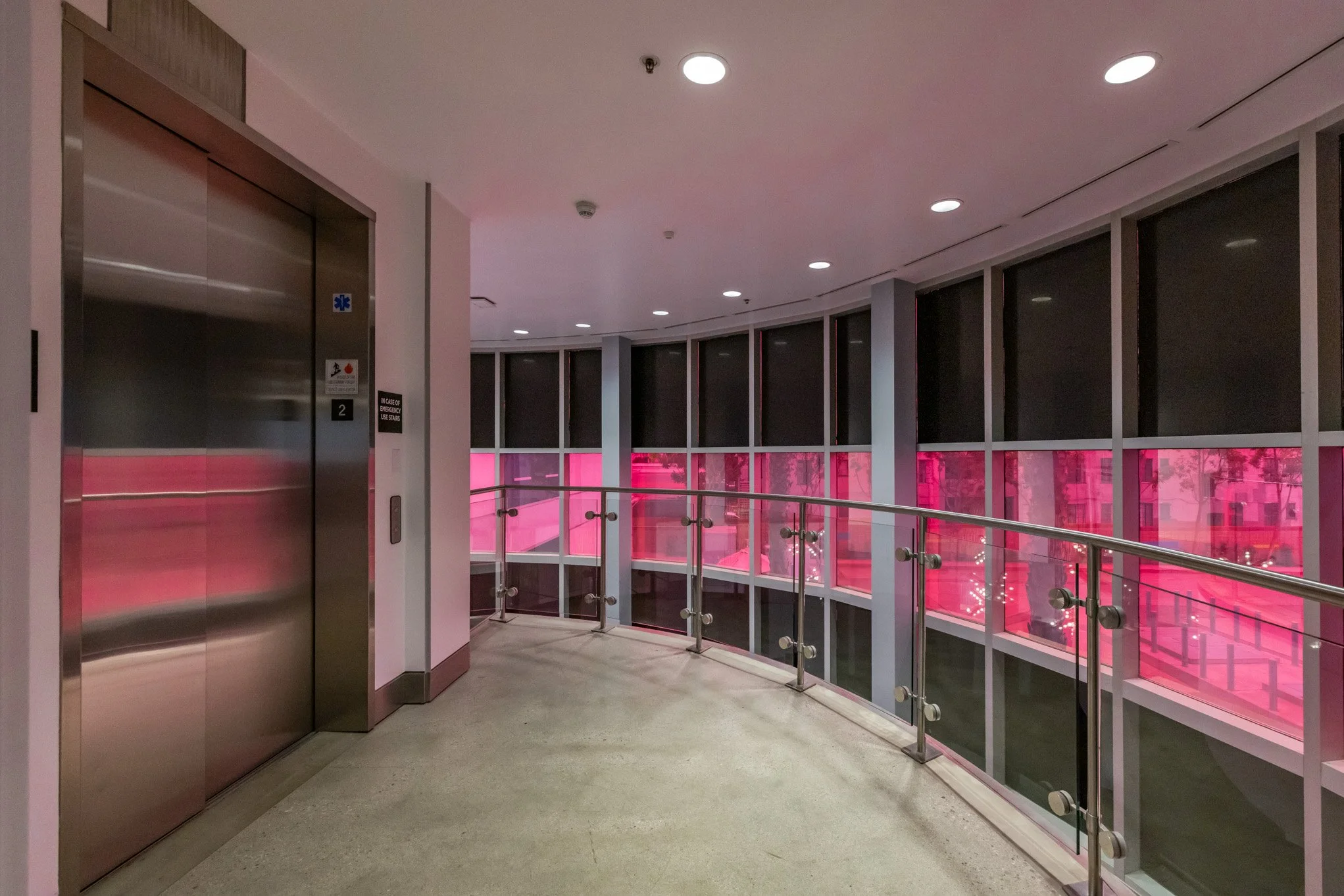 Indoor corridor with an elevator on the left, glass walls with pink tints, and a metal railing along the curved walkway.