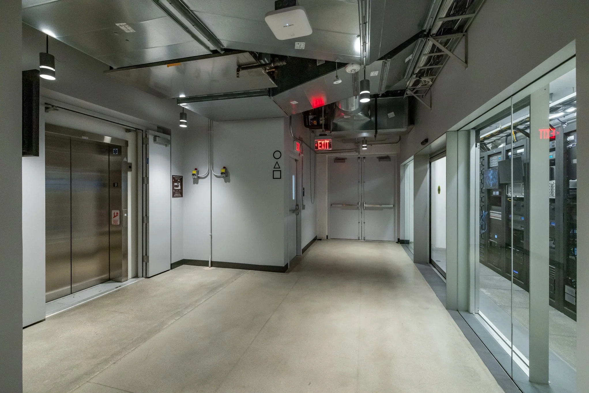 Empty corridor in a modern building, with an elevator on the left, glass doors on the right revealing servers, and a double door at the end, illuminated by ceiling lights and red exit signs.