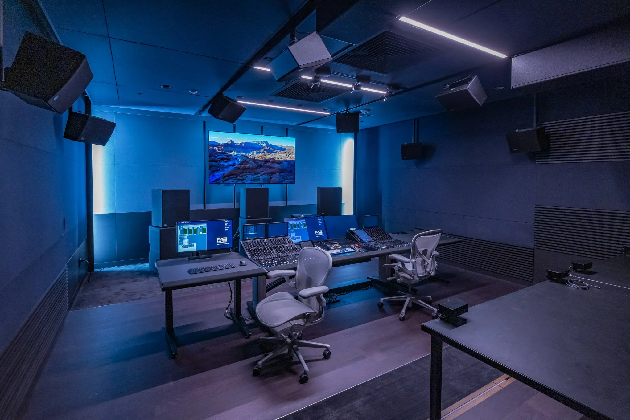 Audio recording studio control room with multiple monitors, sound mixing consoles, and chairs, illuminated by blue lighting.