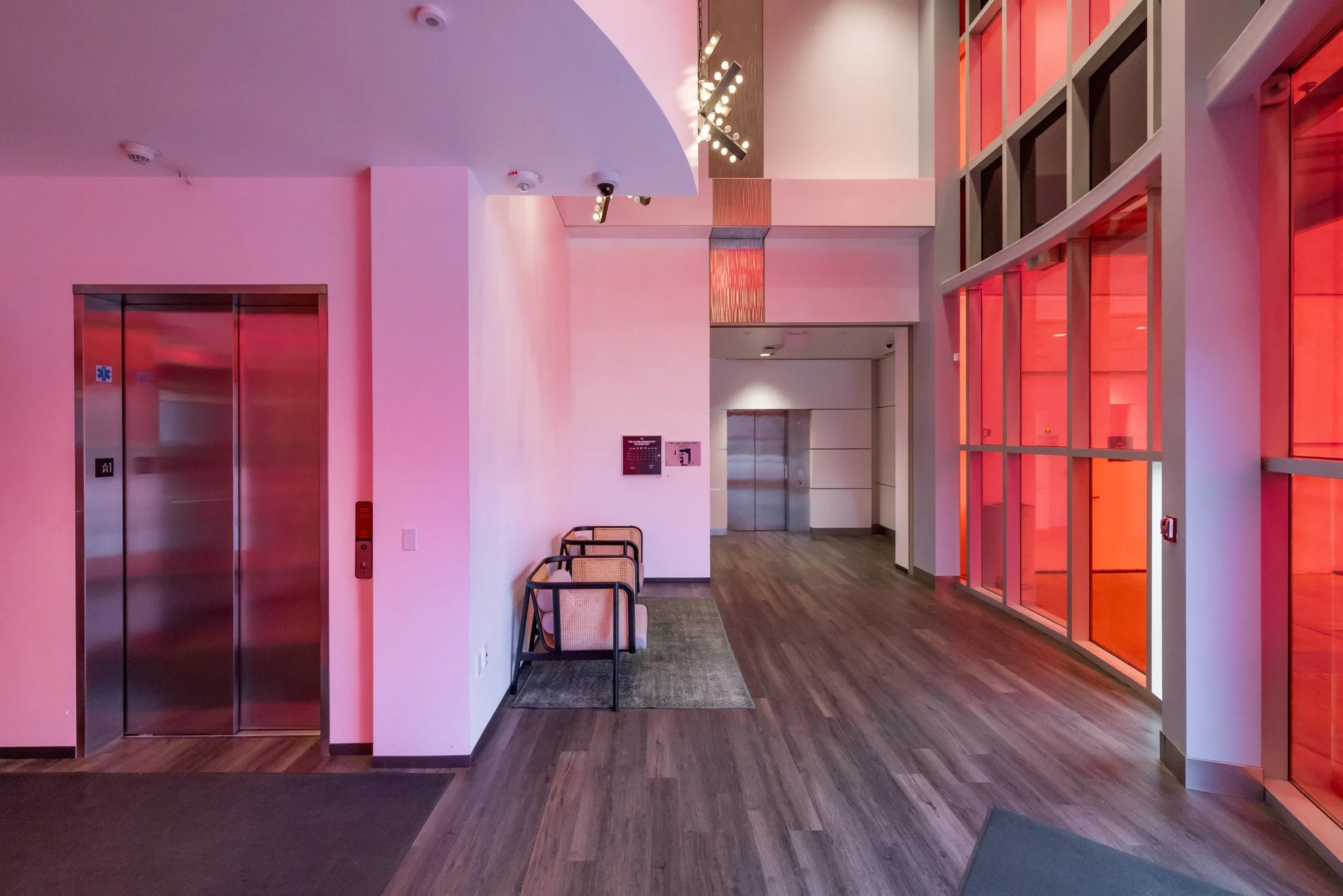 Lobby area with two elevators, wooden floors, a small bench with three chairs, and large windows with red-lit panels.