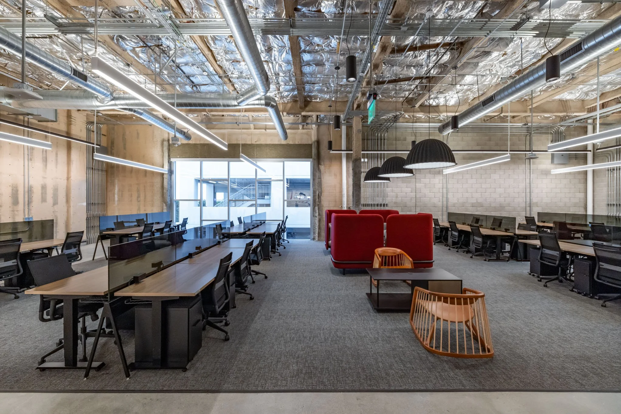 Modern office space with rows of black desks and chairs, some with transparent partitions, exposed ceiling pipes, and a large window at the far end. There are red and orange seating areas with pendant lighting.