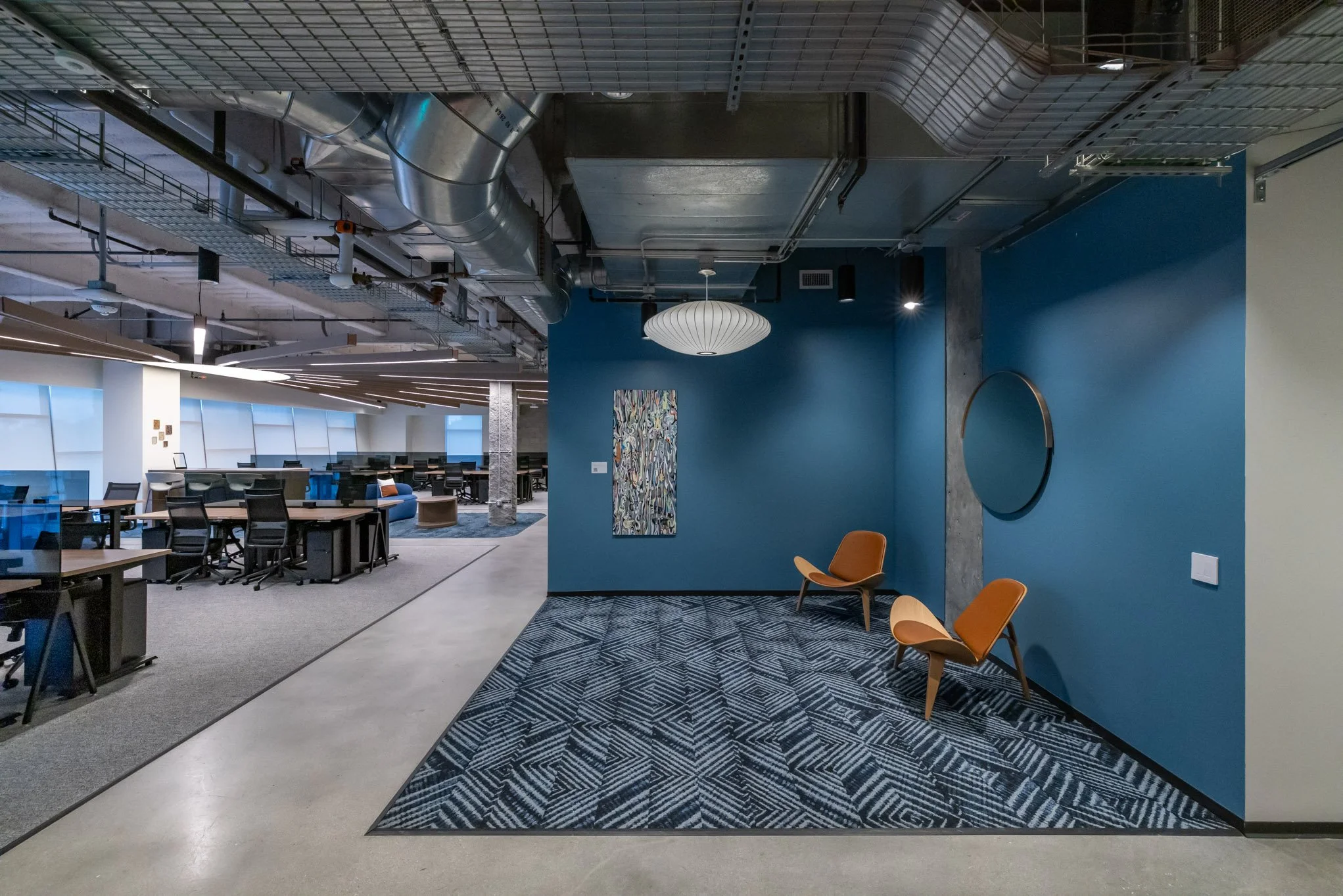 Modern office interior with a small seating area featuring two orange chairs, a blue accent wall with artwork, and an open workspace with desks and chairs in the background.