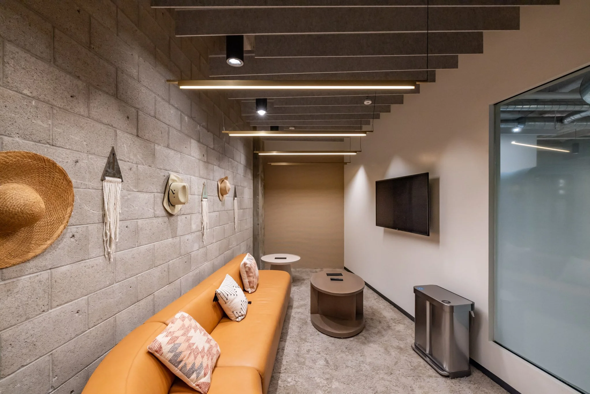 Modern lounge area with a tan leather sofa, decorative pillows, a gray coffee table, a wall-mounted television, a textured gray concrete wall with hanging hats and wall decor, and ceiling lights.